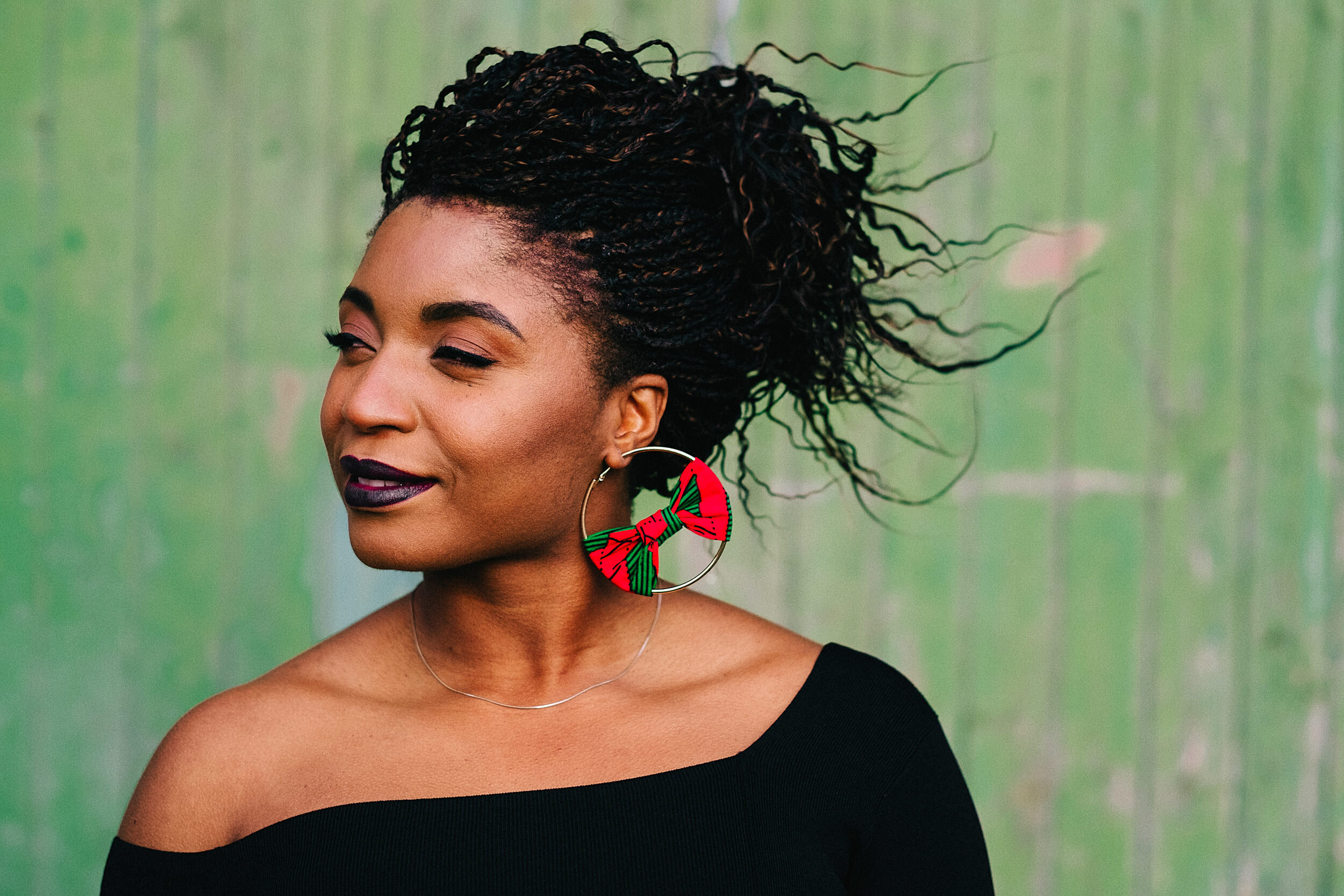 Close-up of a woman with dark curly hair styled in twists, wearing a black off-the-shoulder top, colorful earrings with a red and green scarf pattern, and smiling slightly with a blurred green background.