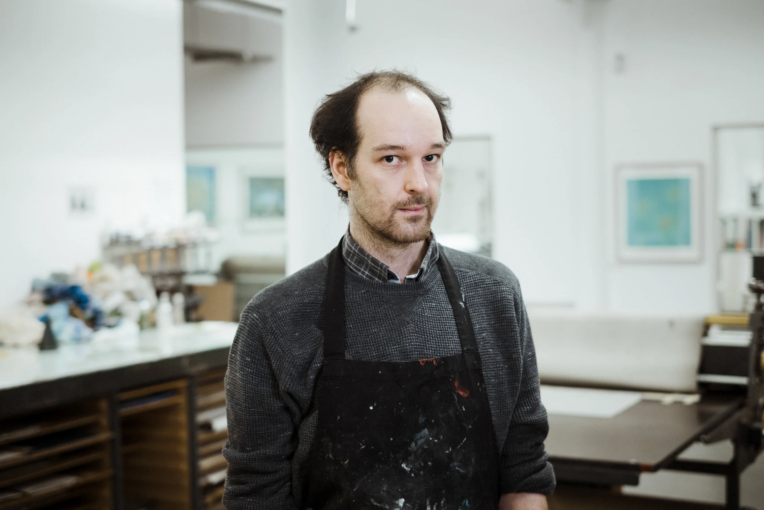 A man with a receding hairline and beard, wearing a black apron and plaid shirt, standing in a well-lit art studio or workshop with tables, shelves, and art supplies in the background.