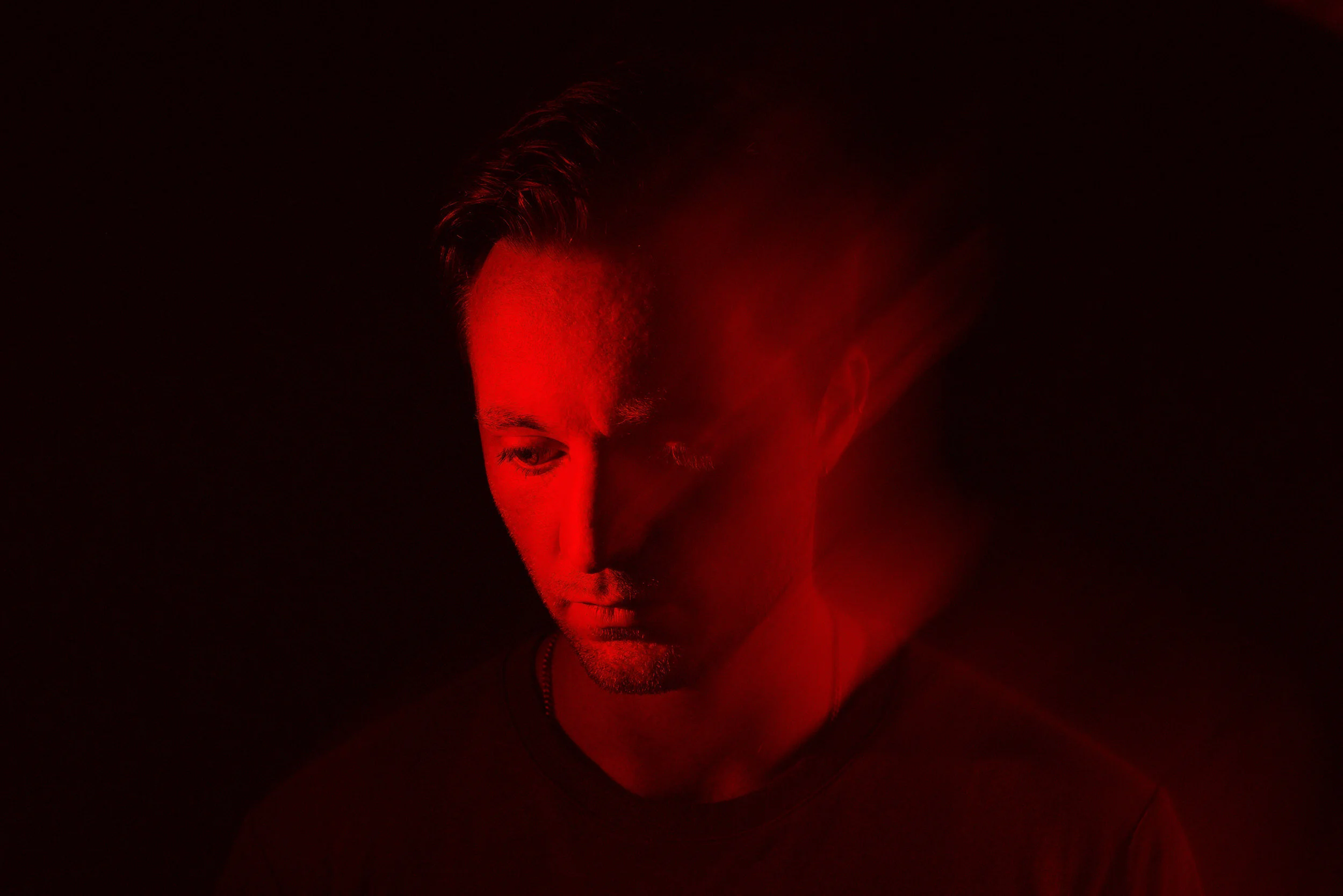 A man with short hair looking downward, illuminated by red lighting against a dark background.