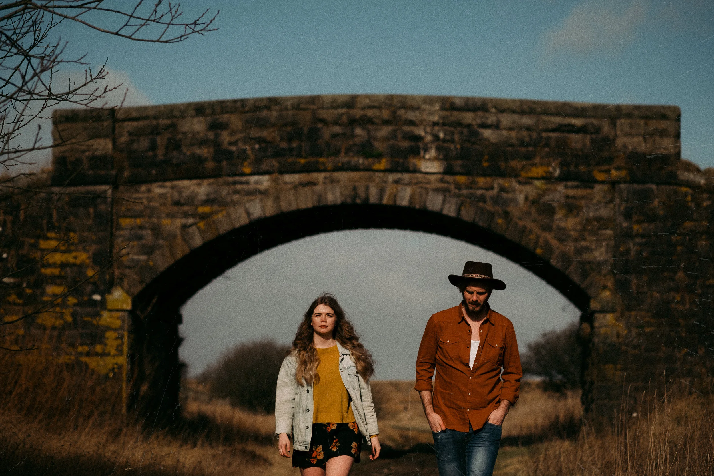 Two people, a woman with long wavy hair wearing a beige jacket and a yellow shirt, and a man with facial hair wearing a brown jacket and a wide-brimmed hat, walking together under an old stone bridge in a rural area during dusk.