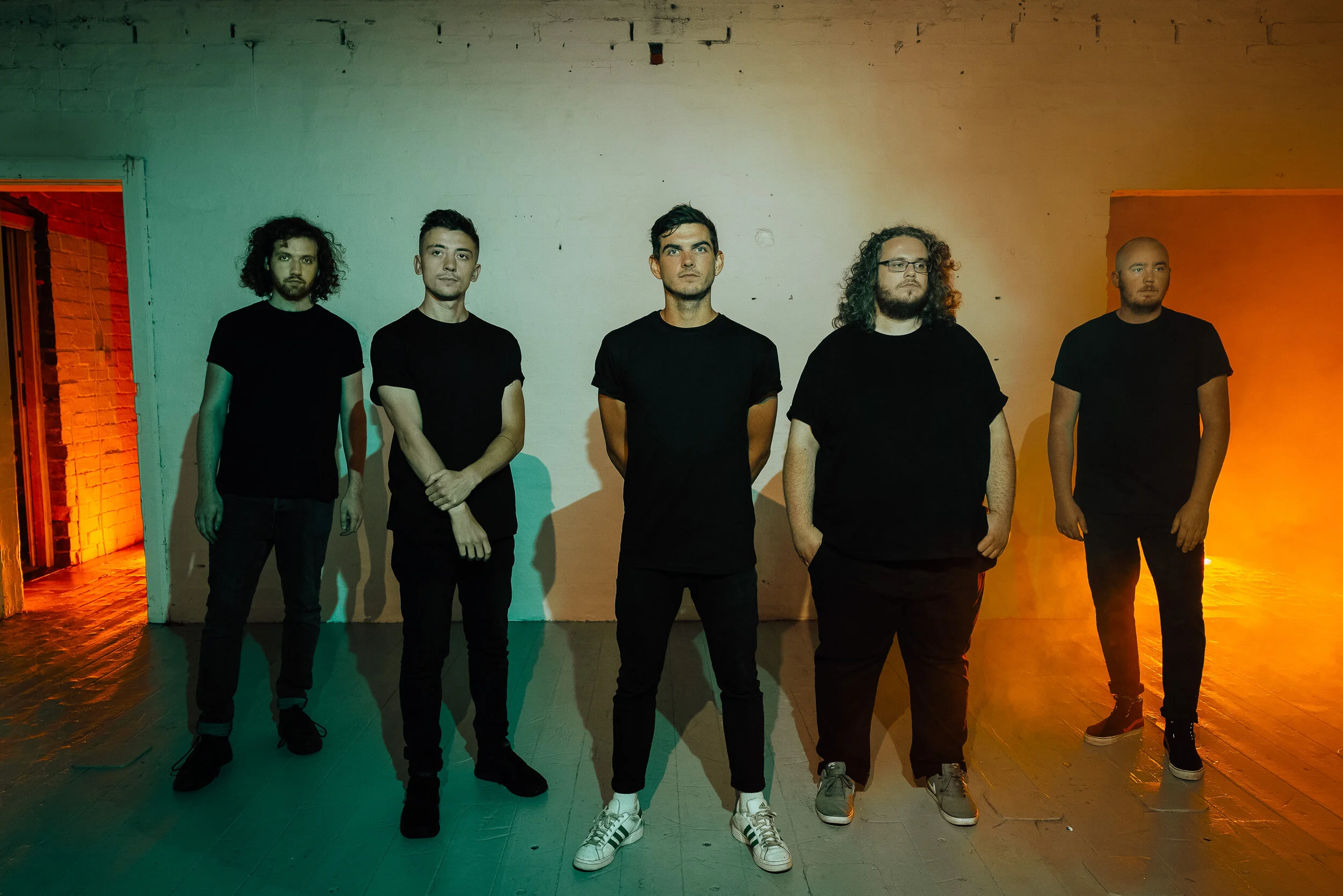 A group of five men standing indoors against a wall illuminated with colored lights, all dressed in black t-shirts and dark pants.
