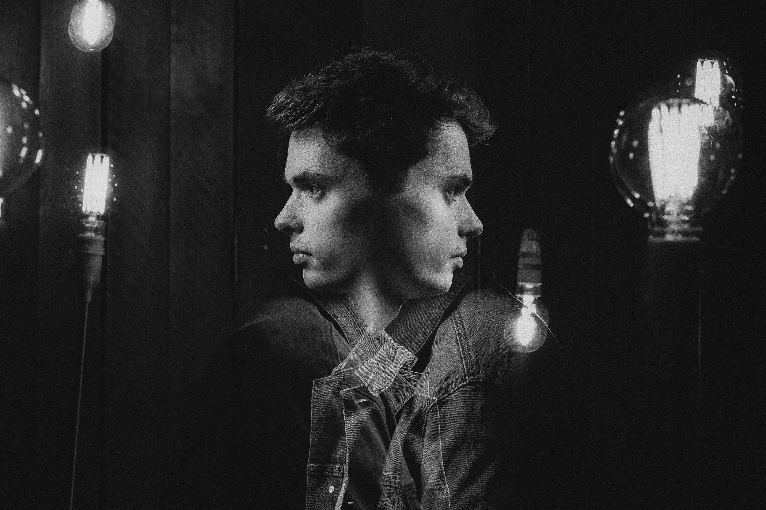 A black and white photo of a person with two faces joined at the center, facing opposite directions. The person is in a dark room with illuminated light bulbs on either side.