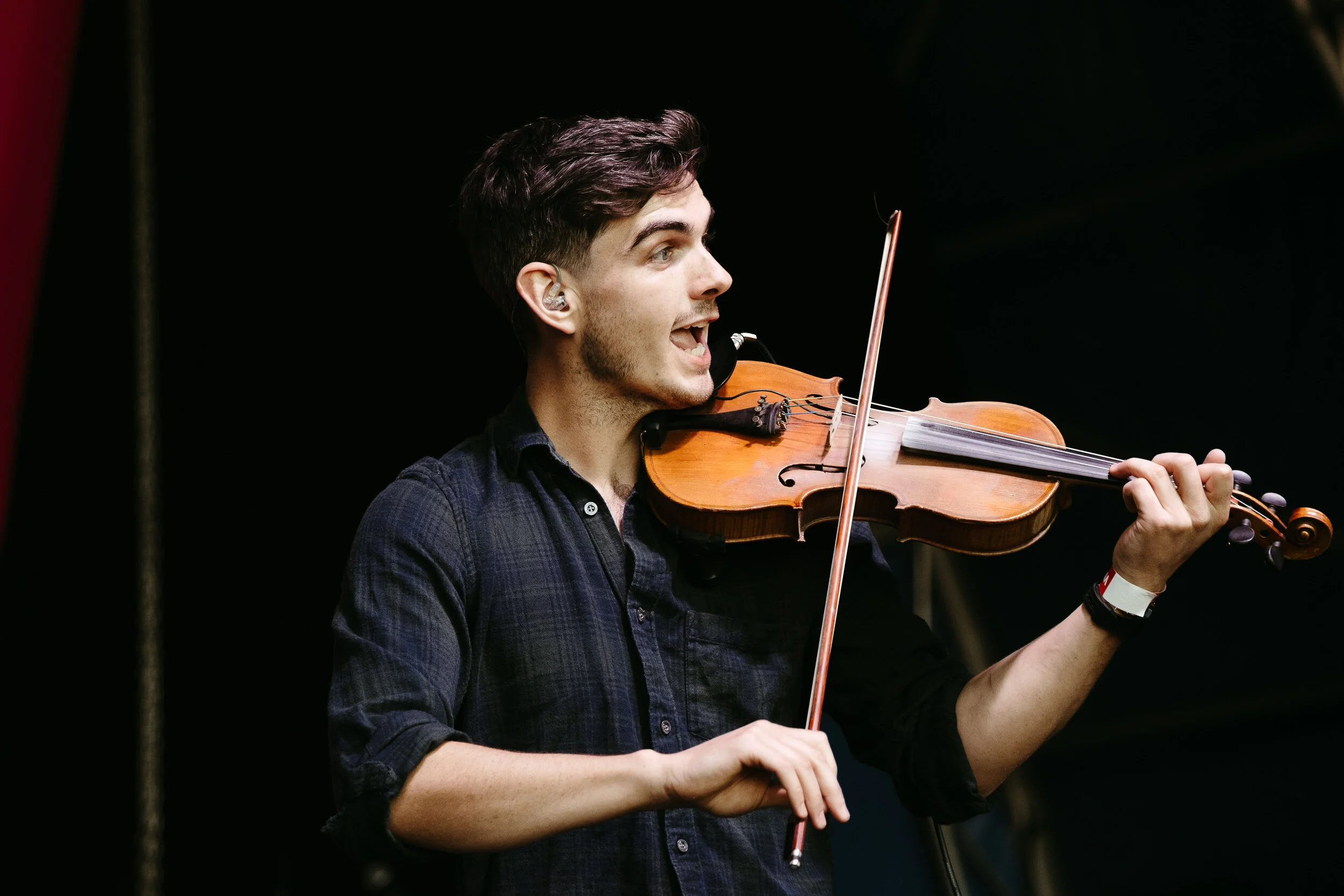 A young man playing a wooden violin passionately, with a focused expression and wearing in-ear monitors, against a dark background.