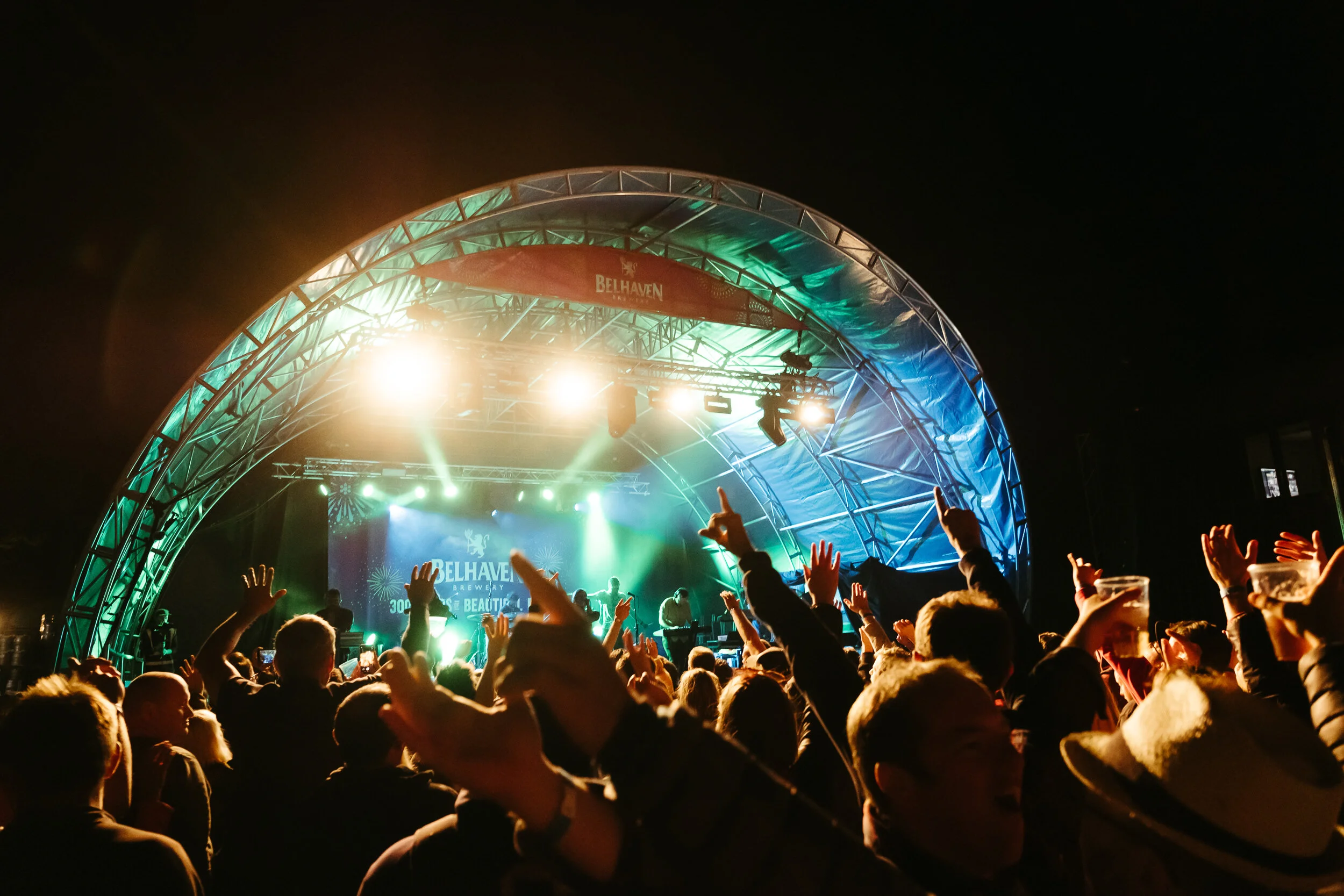 Crowd enjoying a concert at night with a stage labeled Belhaven Brewery, illuminated by colorful lights.