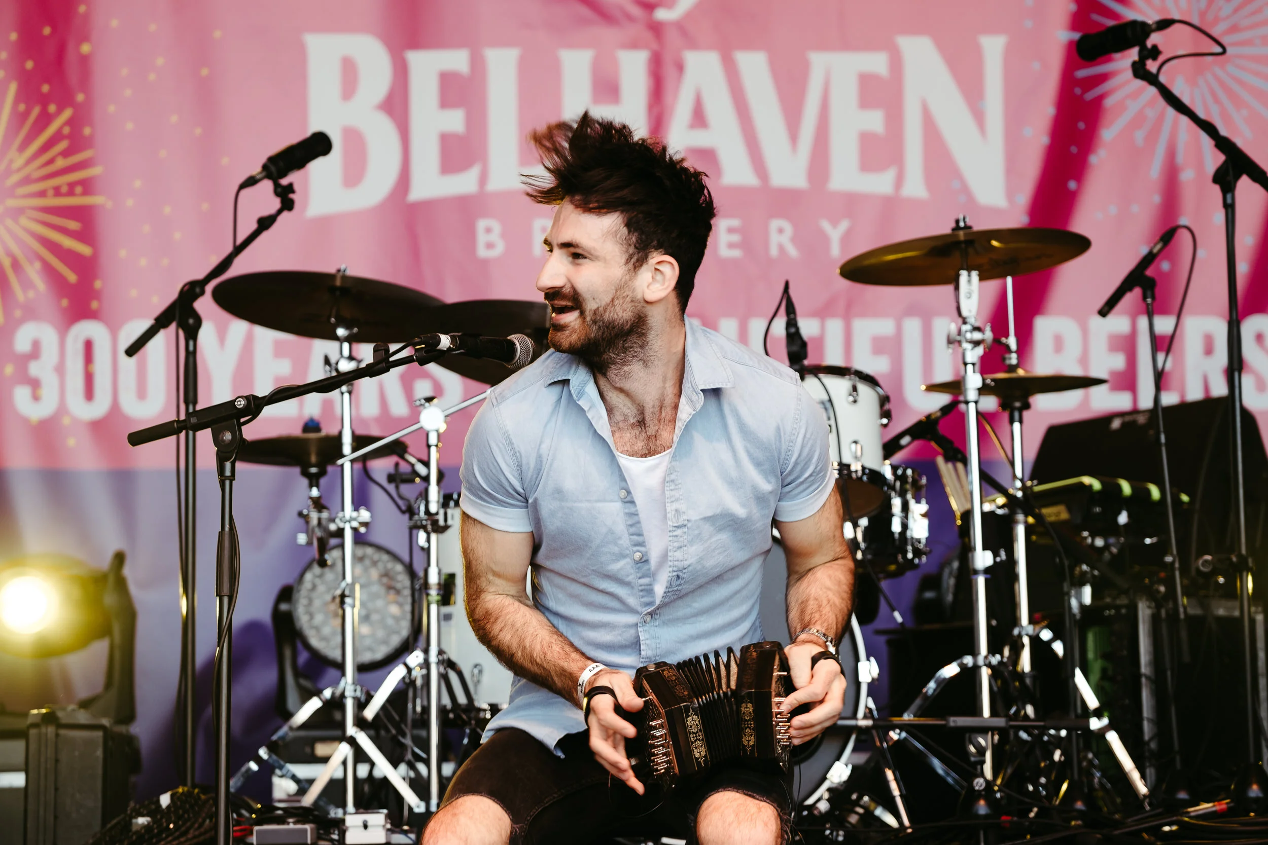 A man with dark hair and a beard is playing a musical instrument at a live performance. He is smiling and wearing a light blue shirt. There is a drum set and microphones in the background, along with a pink backdrop with white text that reads "BELHAV