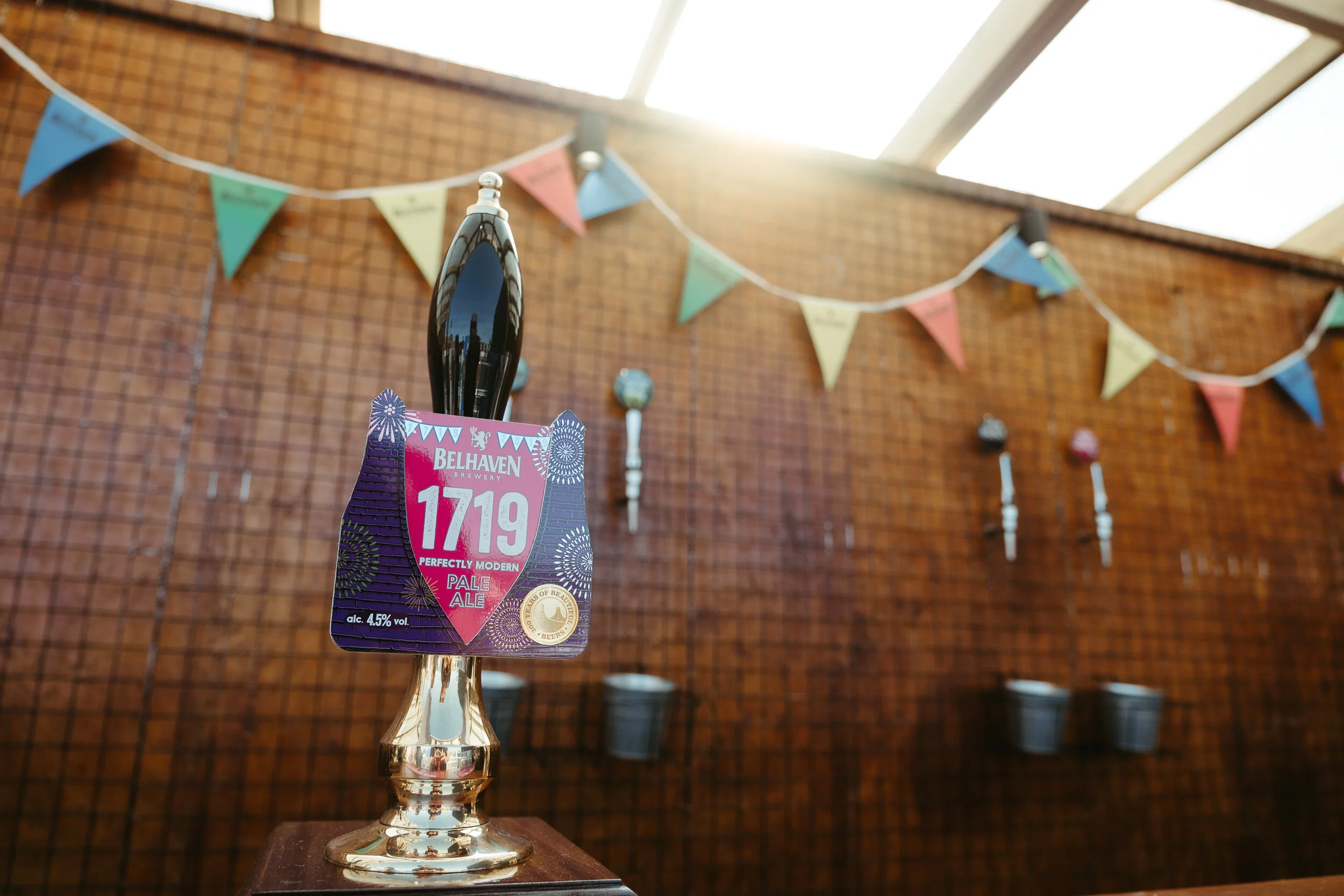 A beer tap handle with a label from Belhaven Brewery, featuring a pink and purple design with fireworks and the words 'Perfectly Modern Pale Ale' and '4.5% vol' against a background of a party or celebration setting with colorful bunting and a brick 
