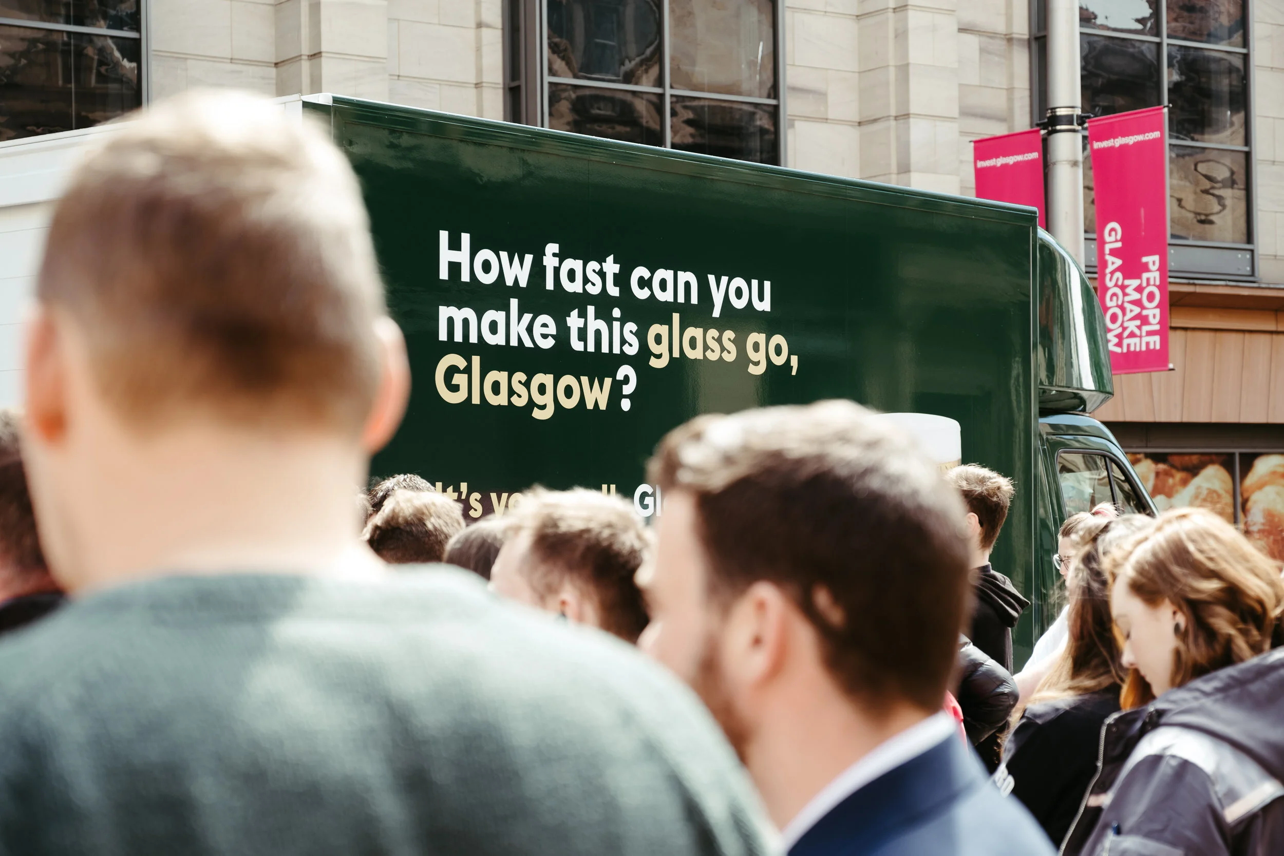 Crowd of people gathered outdoors in front of a green van with white text asking how fast one can make glass go in Glasgow, Scotland.