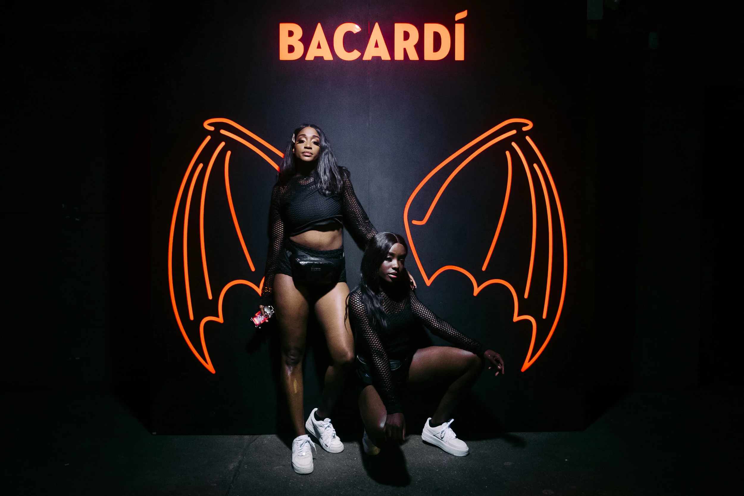 Two women with dark hair and dark skin posing in front of a neon orange bat wing design on a black wall. One woman is standing, holding a phone, and wearing a black mesh top and black shorts. The other woman is sitting, wearing a similar black mesh t