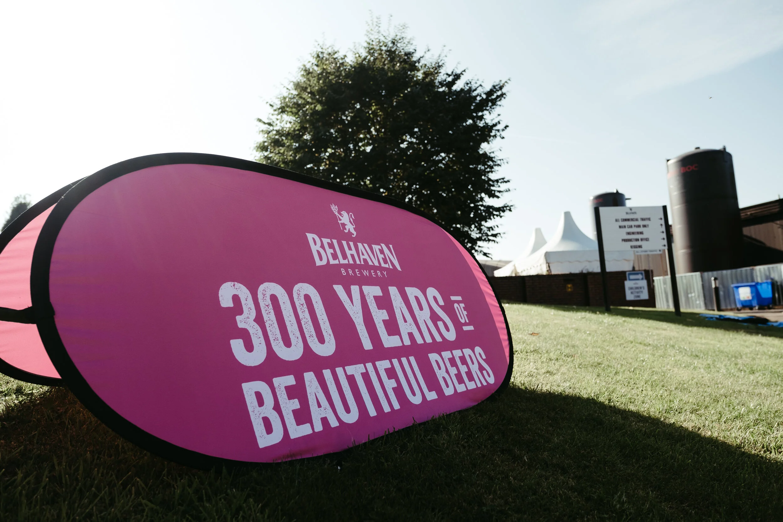 Pink signboard commemorating 30 years of Belhaven Brewery with white text, placed on grass outdoors, with trees, a white tent, and black pipes in the background under a clear sky.