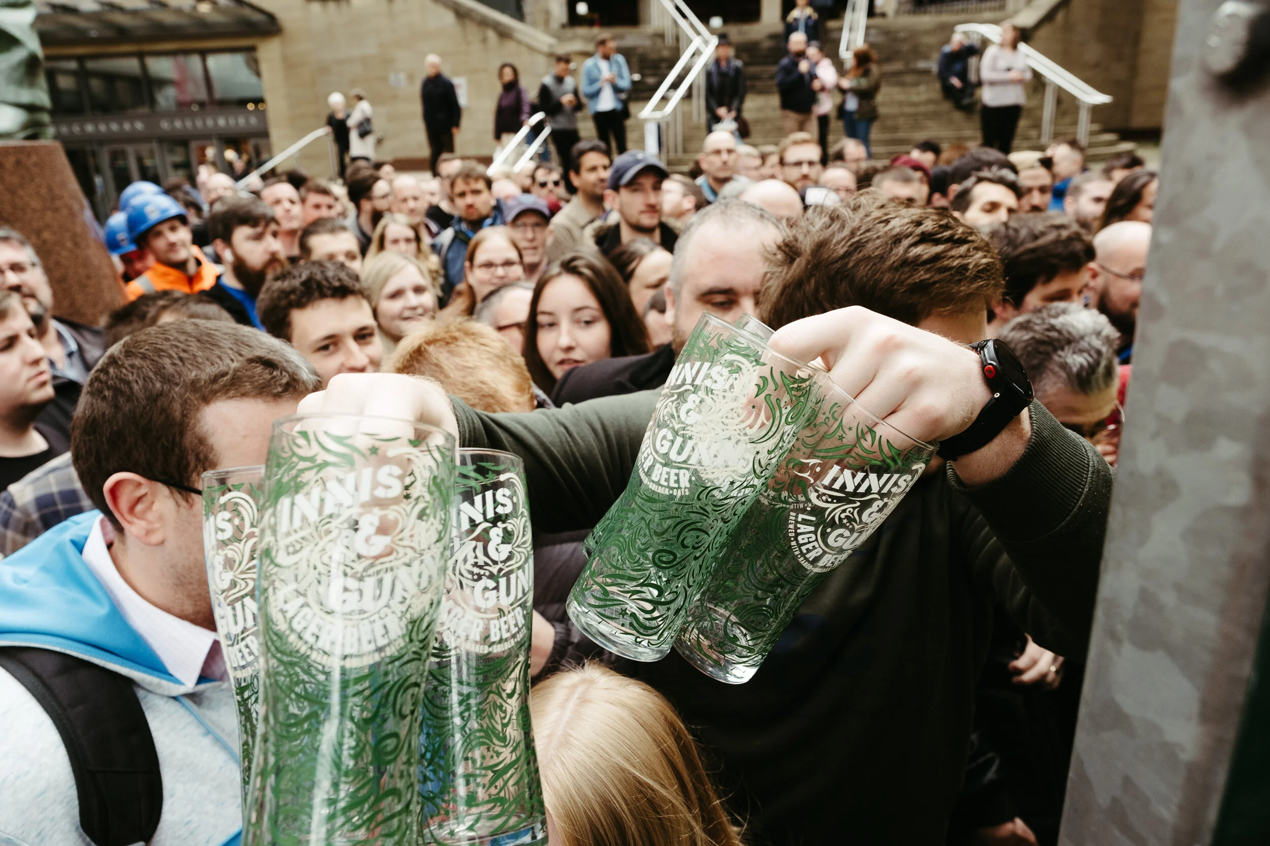 A crowd of people gathered outdoors, raising glasses in a toast. The glasses have a decorative pattern and some text related to beer.