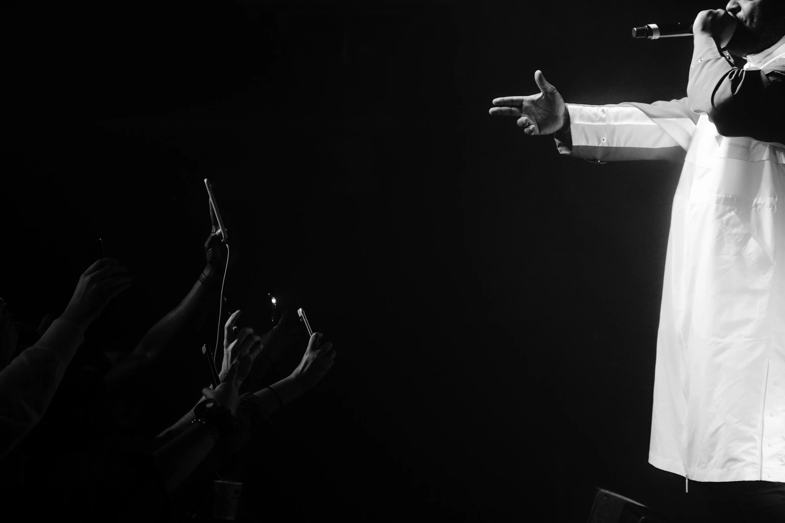A performer on stage extending his hand towards the audience, with multiple fans reaching out and holding smartphones. The image is in black and white, capturing the moment of connection during a performance.