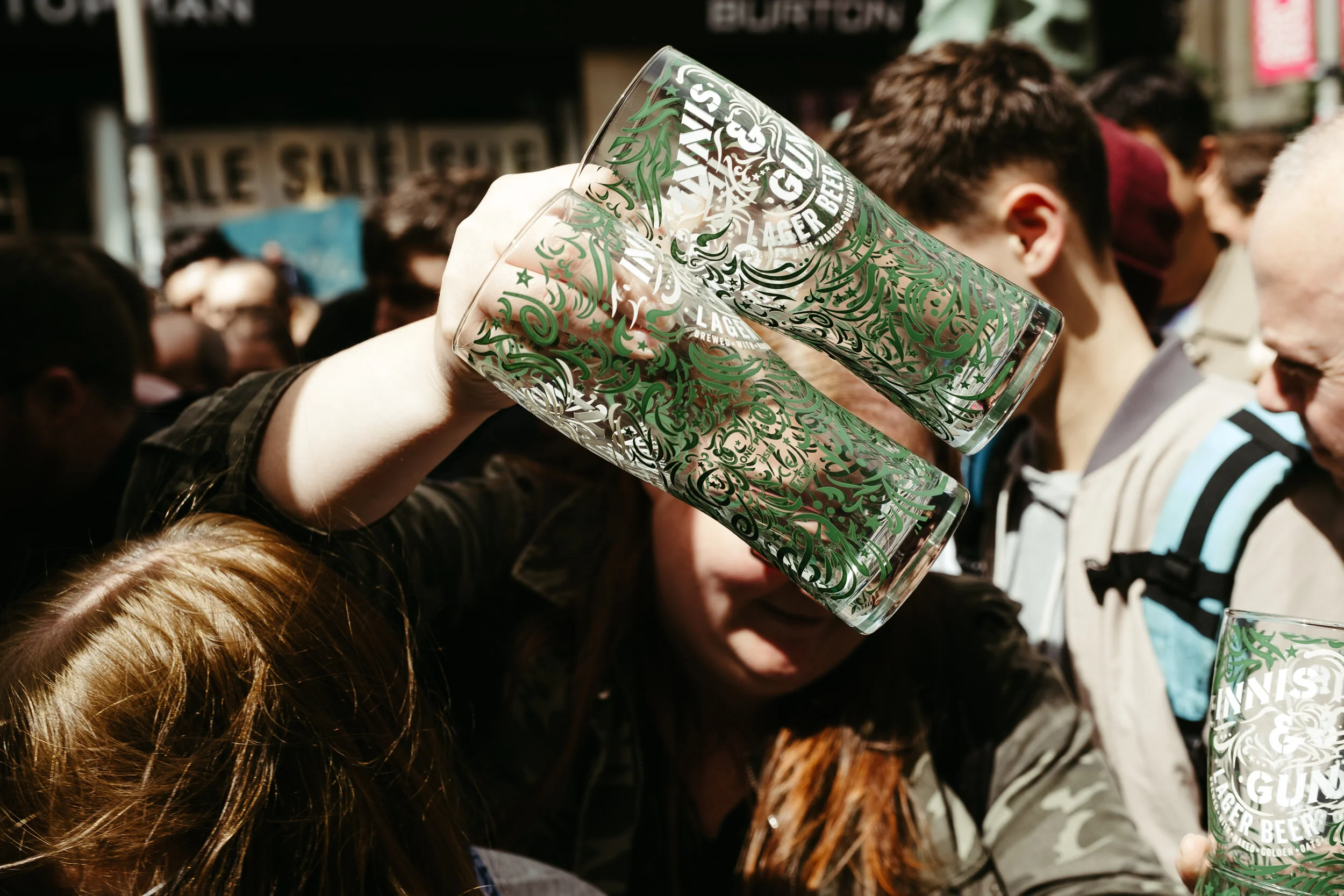 A glass with green decorative designs being held above a crowd at an outdoor event, featuring people standing closely together.