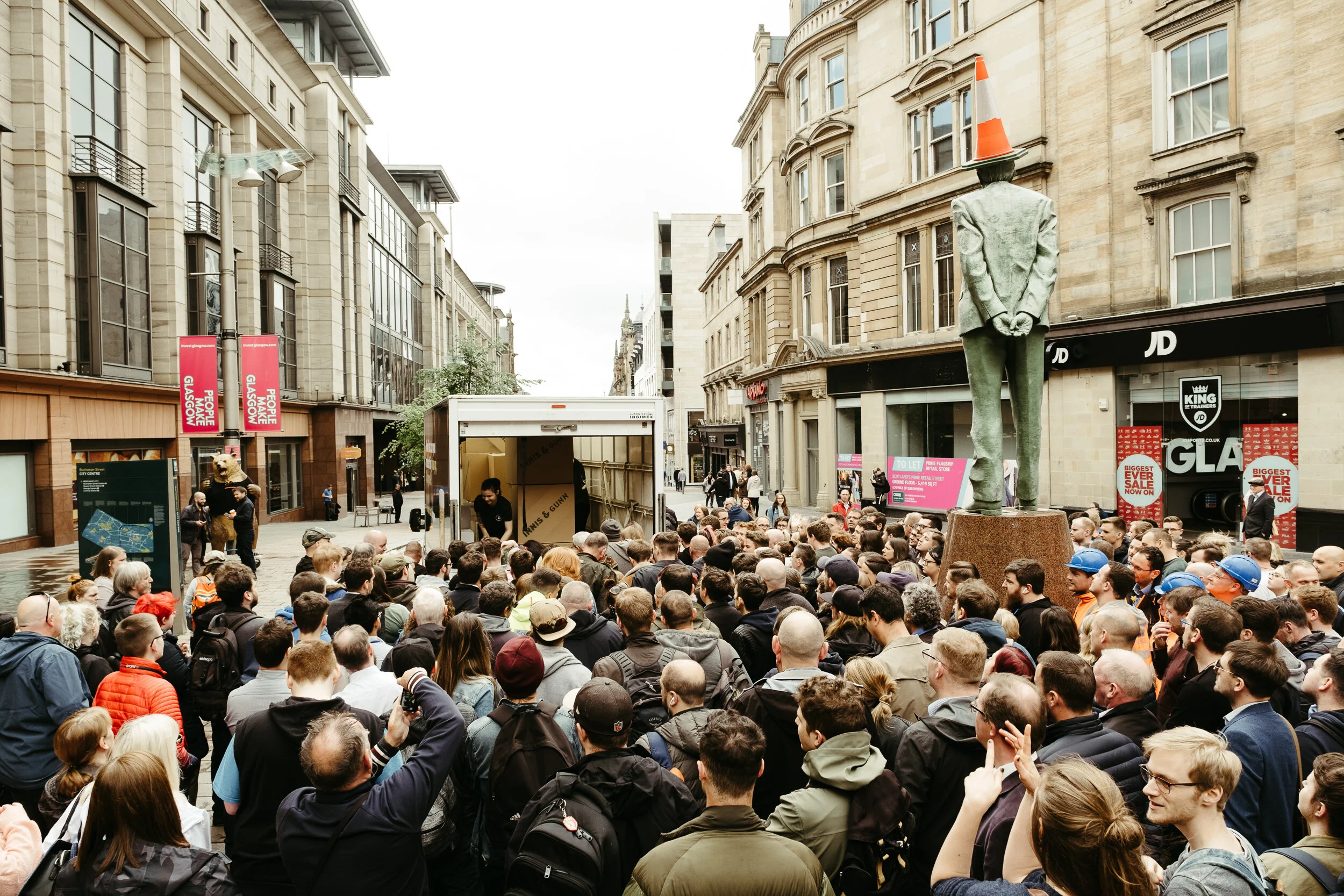 Crowd of people gathered in a city street under a cloudy sky, with buildings on both sides and a statue with a traffic cone on its head in the street.