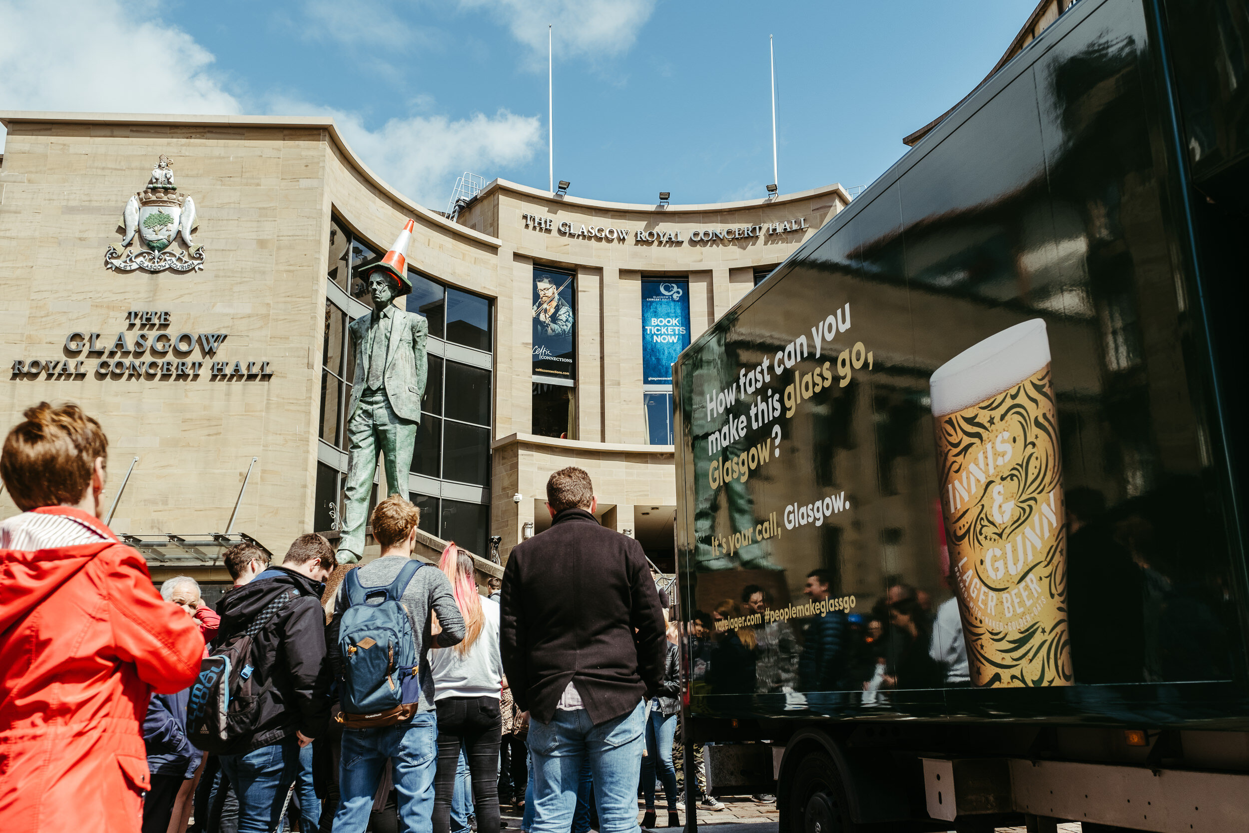 Crowd of people standing outside The Glasgow Royal Concert Hall in Glasgow, Scotland. The building has a crest on its wall, and statues are visible near the entrance. A large black truck with advertisements on the side is in the foreground.