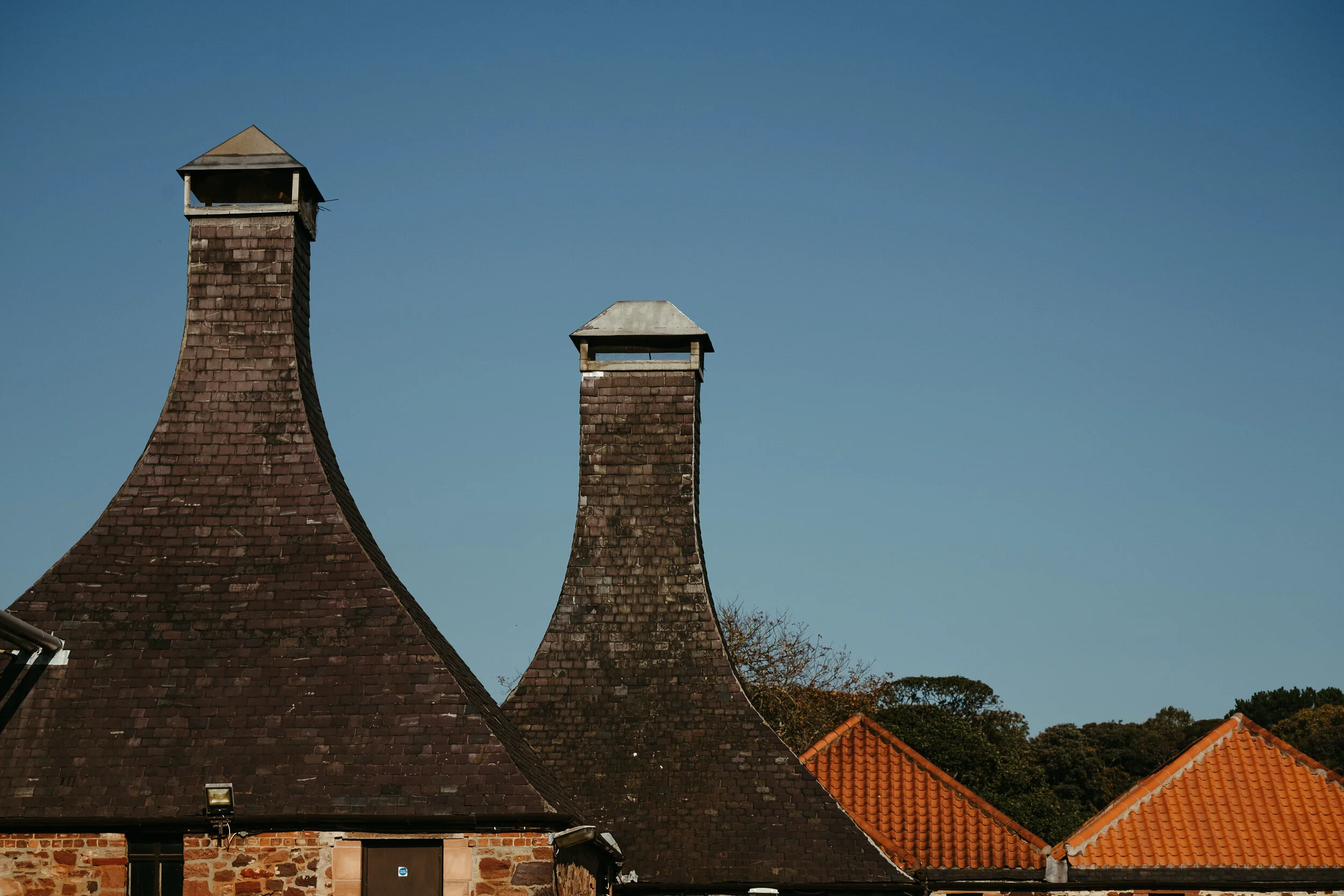 Three brick chimneys with different shapes and roof styles extend above the rooftops of brick buildings; a clear blue sky is in the background.