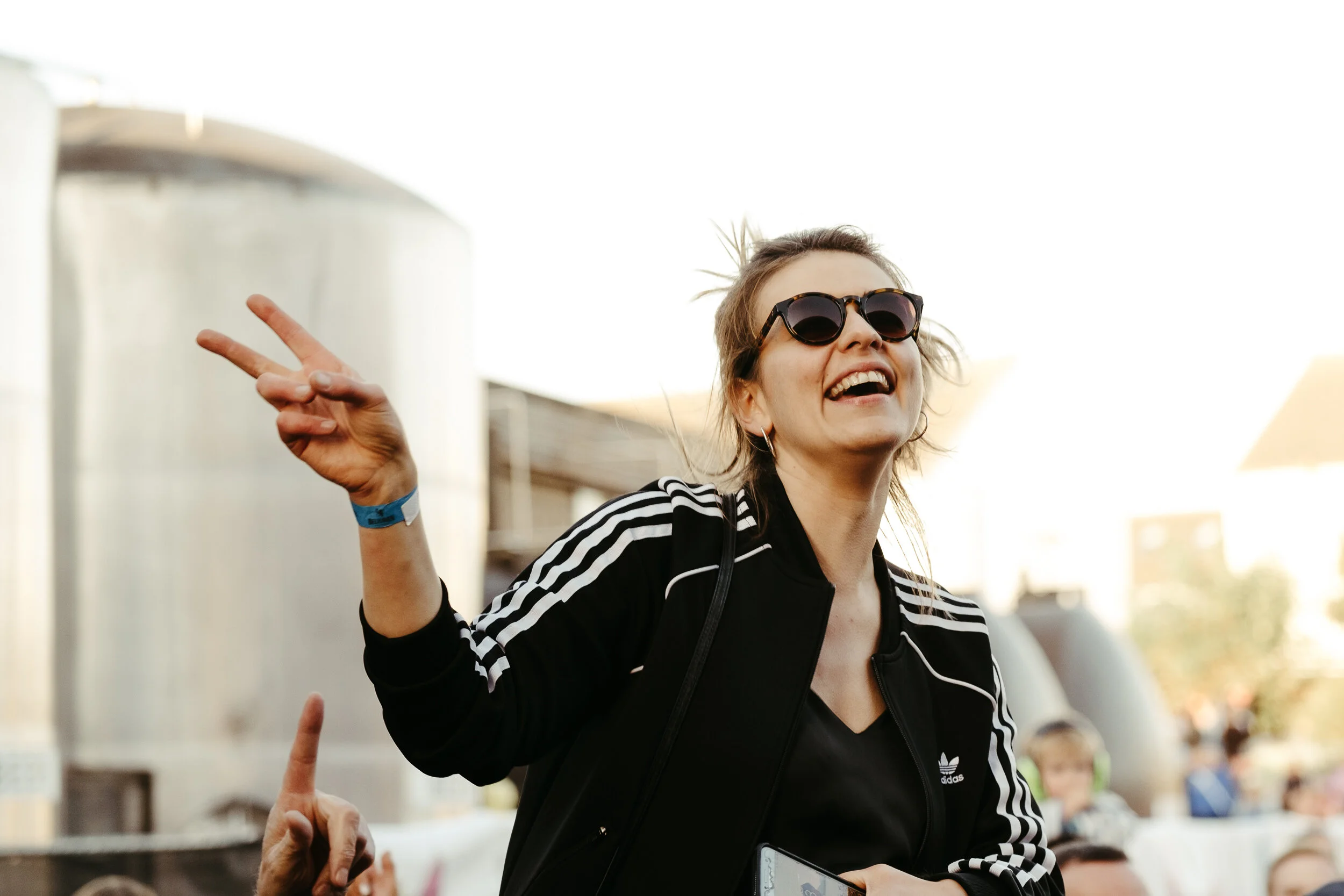 A woman with short hair, wearing sunglasses and a black Adidas jacket, smiling and making a peace sign with her fingers in an outdoor setting with other people around.