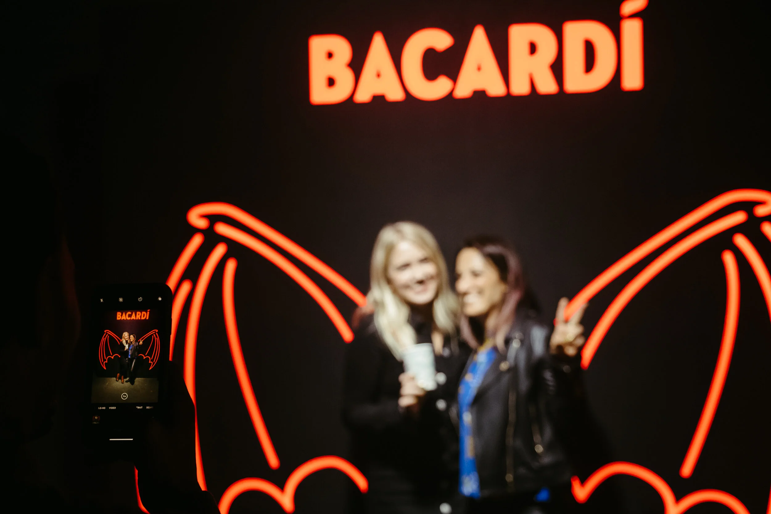 Two women taking a selfie in front of a neon sign that says Bacardi with bat wings underneath.