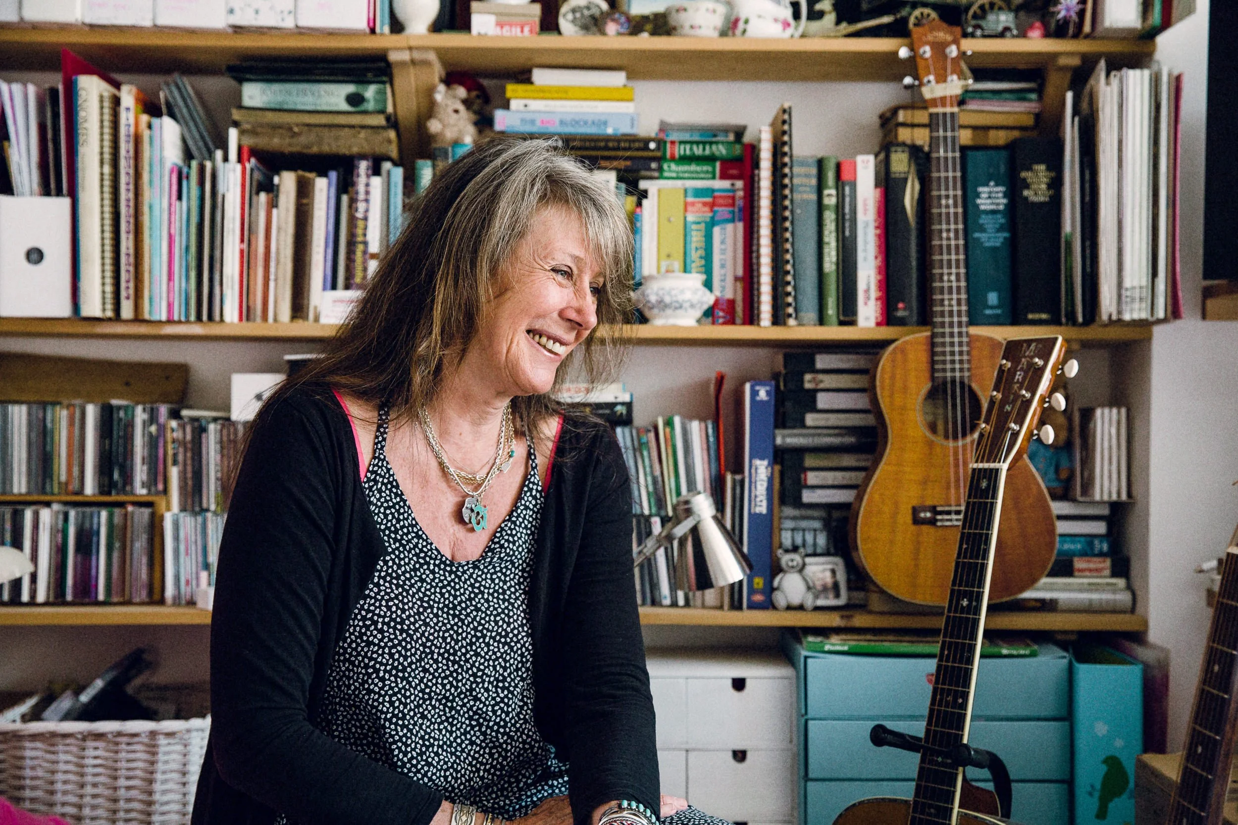 A woman with long gray hair smiling while sitting at a desk in a room with a bookshelf full of colorful books and a guitar standing on a stand. The room has a cozy, lived-in feel with various decorative items.