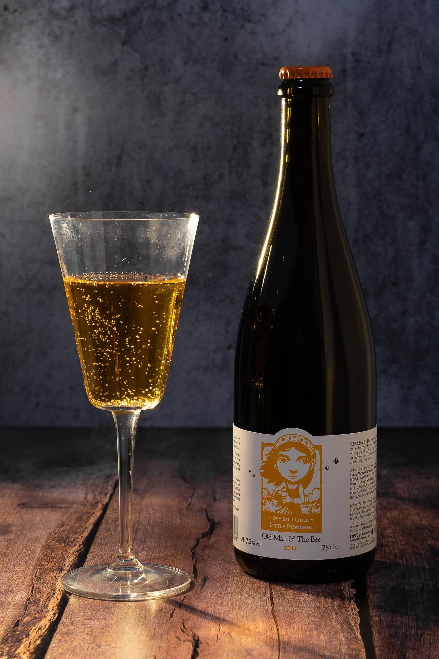A glass of sparkling apple cider next to a dark bottle with a label featuring a cartoon girl and the brand name 'Old Man & The Bee' on a wooden surface with a dark textured background.