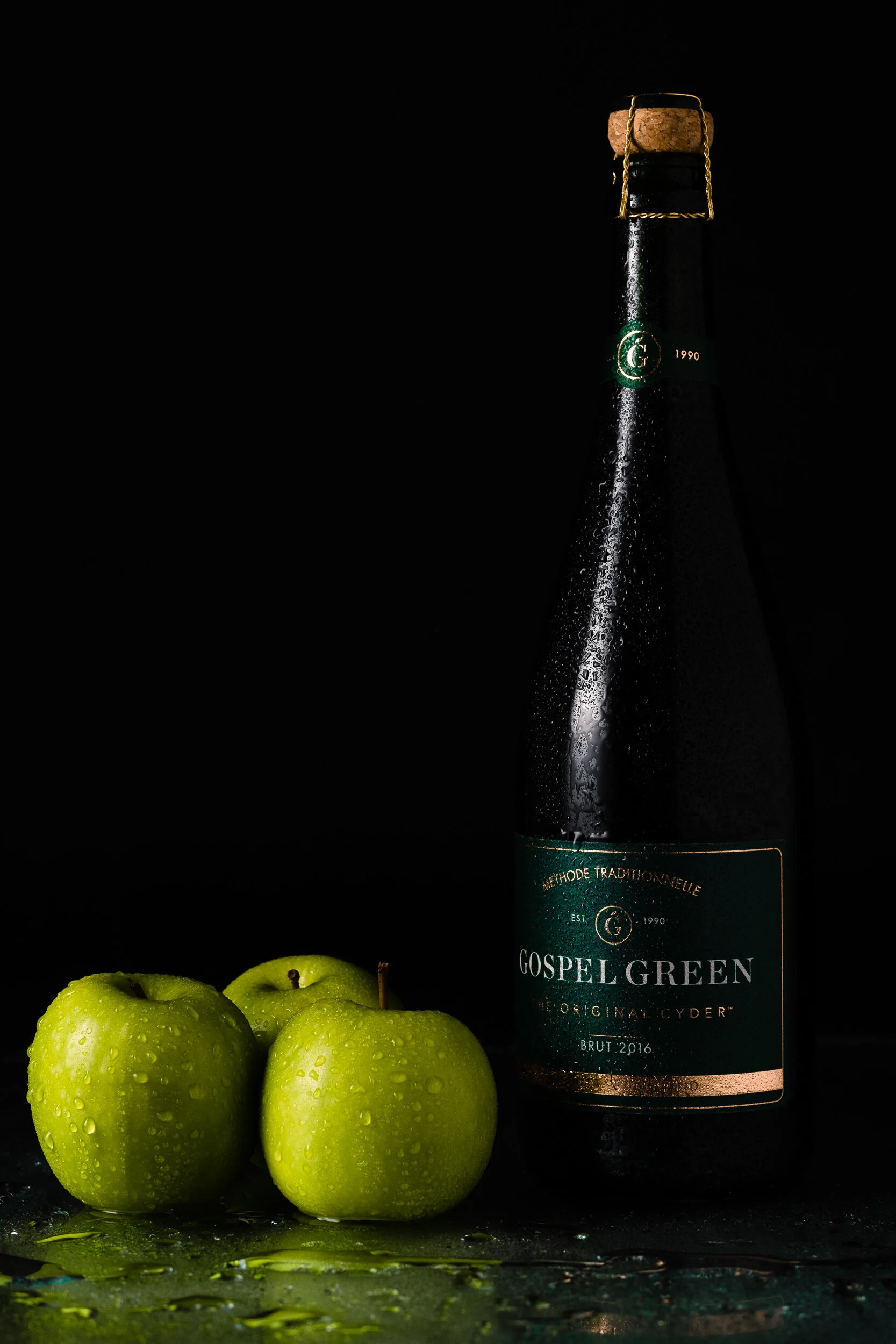 Three green apples with water droplets next to a dark green bottle of Gospel Green cider with a cork and a gold wire cage, set against a black background with water droplets and spilled liquid surface.