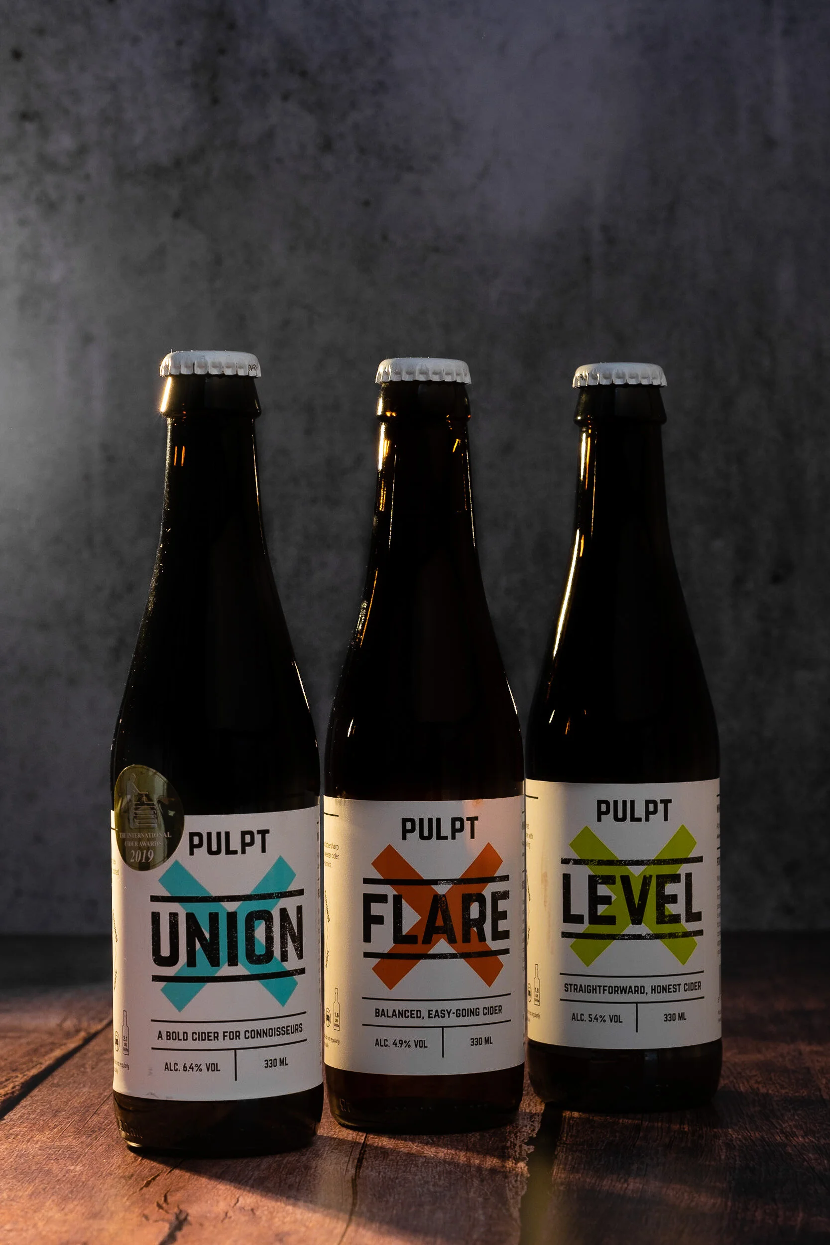 Three bottles of cider standing side by side on a wooden surface, with dark textured background. Each bottle has a white label with bold black, colorful text, and metallic caps. The labels read 'Union,' 'Flare,' and 'Level,' respectively, with additi