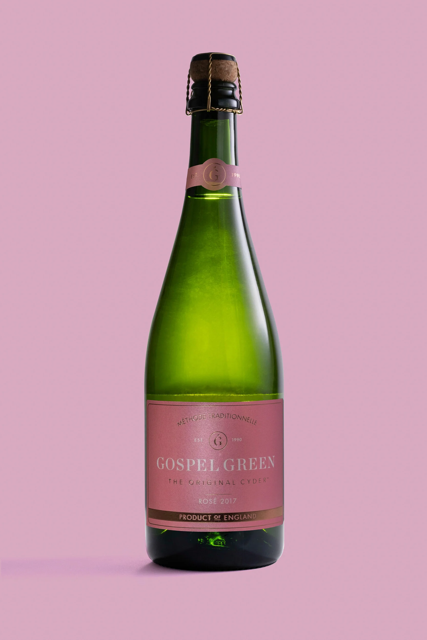 A green glass bottle with a pink label and a cork stopper, set against a pink background.