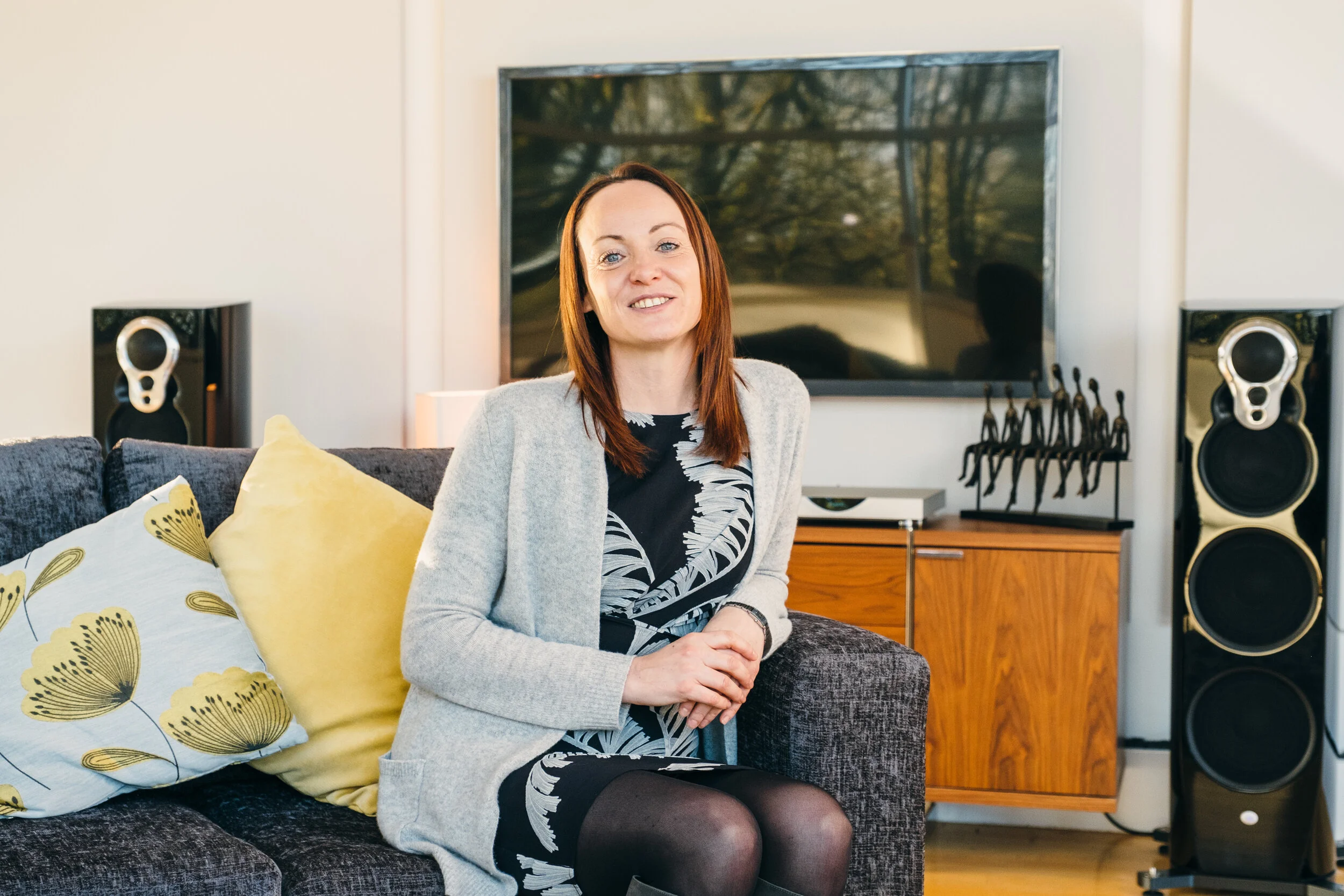 A woman with red hair sitting on a dark gray sofa in a living room, wearing a black and white dress with a gray cardigan, and black tights, smiling at the camera.