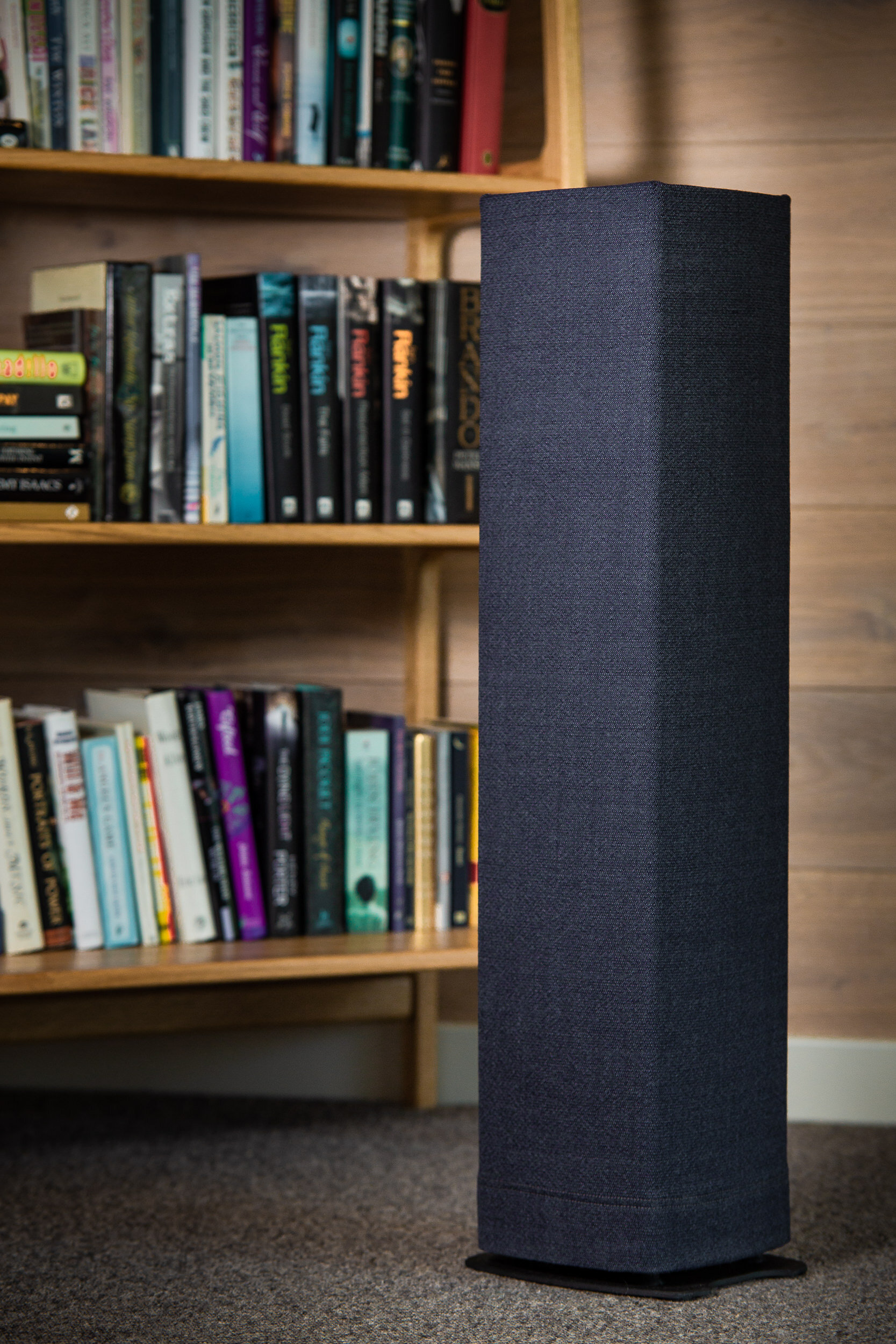 A black speaker standing on the floor in front of a wooden bookshelf filled with various books.
