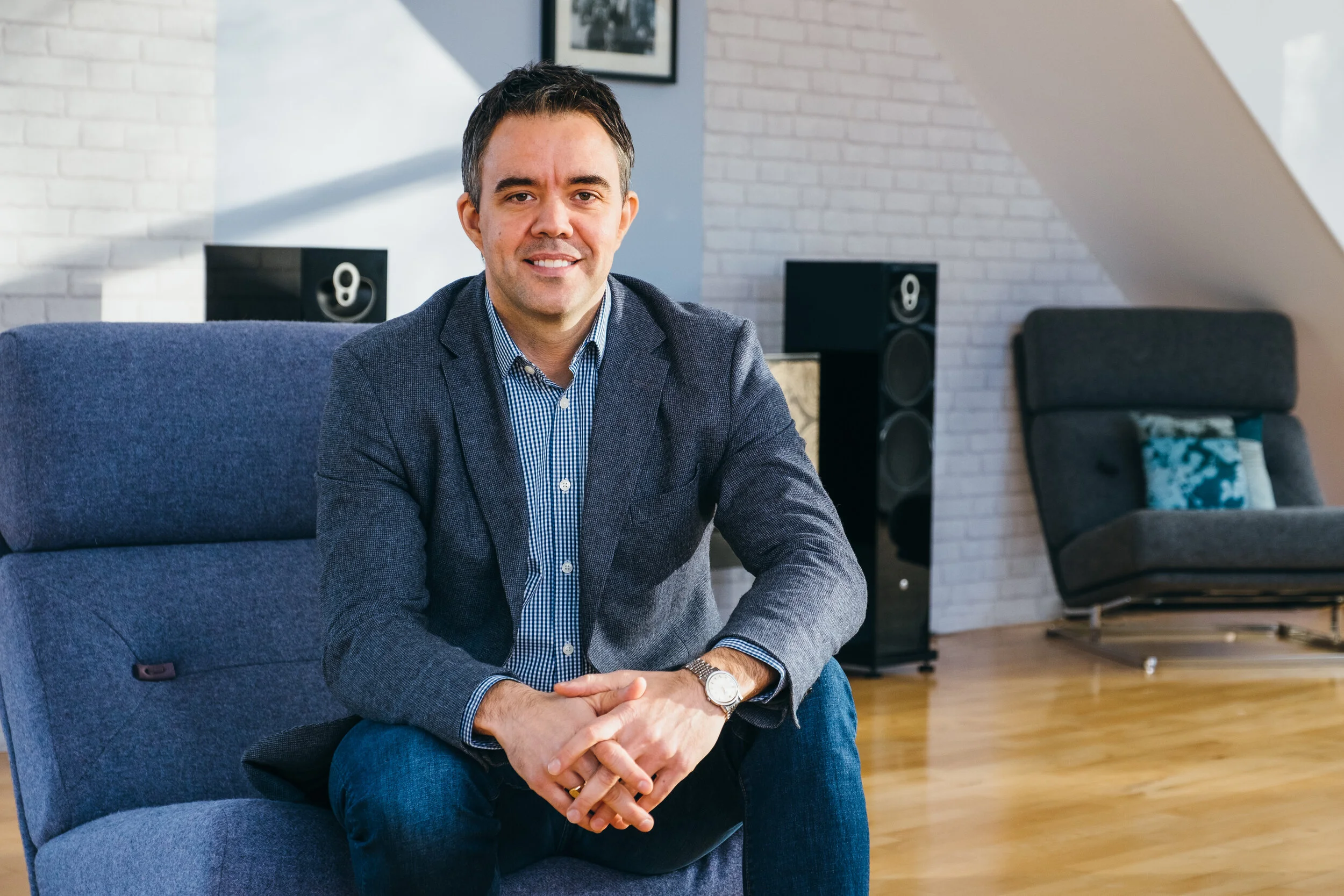 A man sitting on a blue couch in a modern living room, smiling at the camera, with brick walls, framed art, speakers, and a chair in the background.
