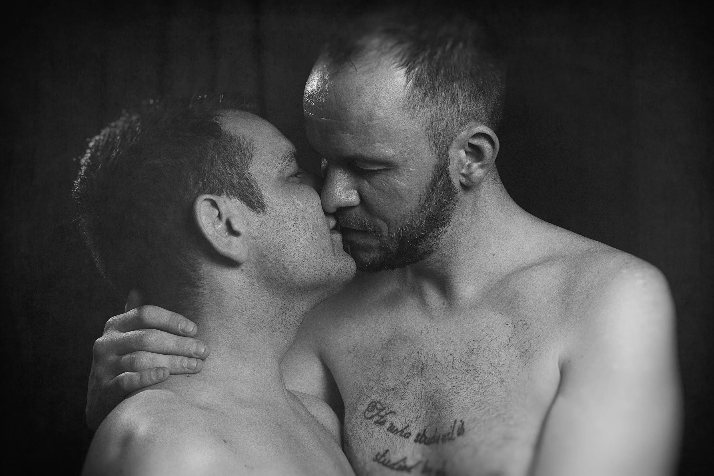 Two shirtless men, one with short dark hair and the other with short light hair and a beard, are engaged in a close, intimate embrace and kiss against a dark background.