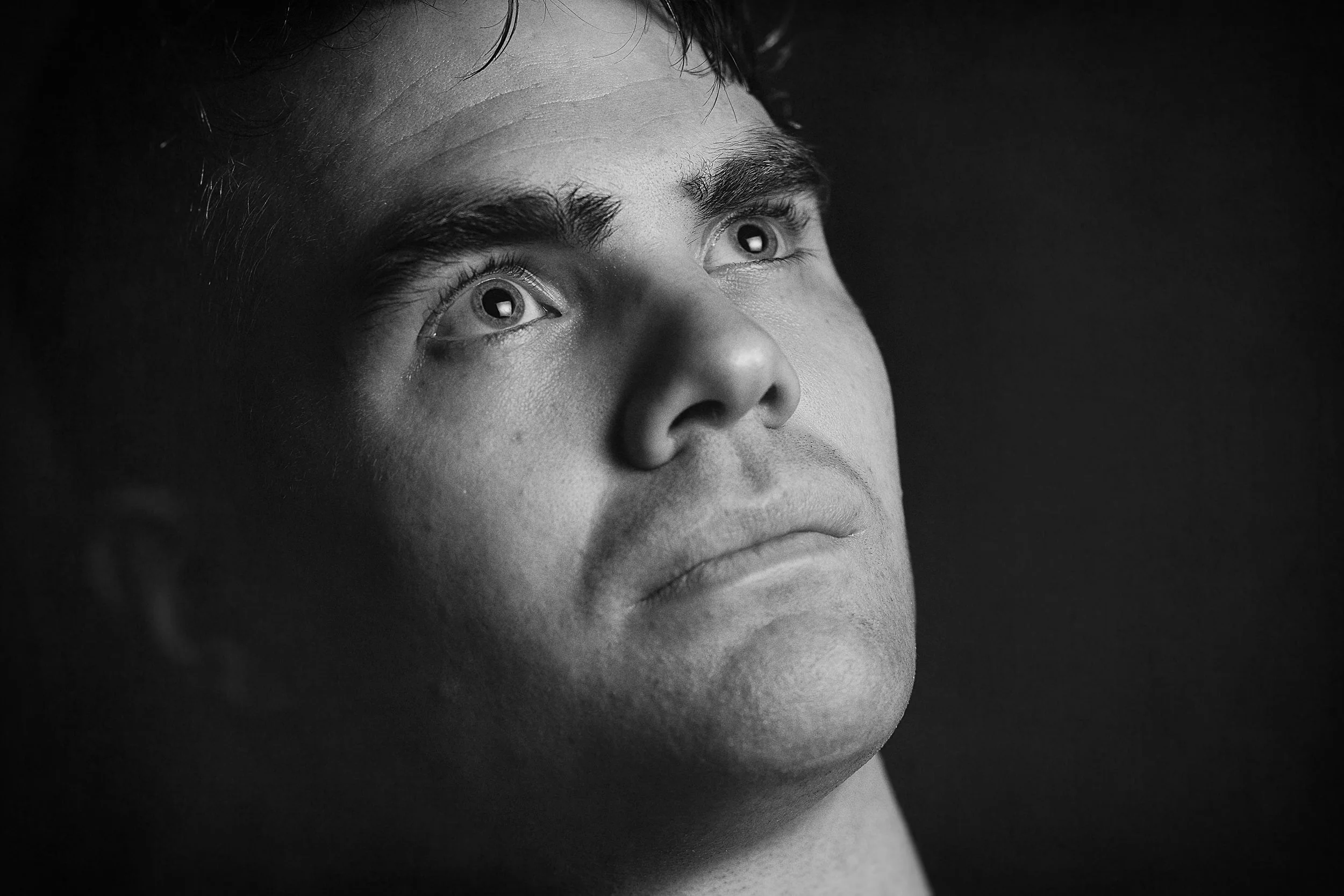 Close-up black and white portrait of a young man looking upwards, showing his face from the nose up with a dark background.