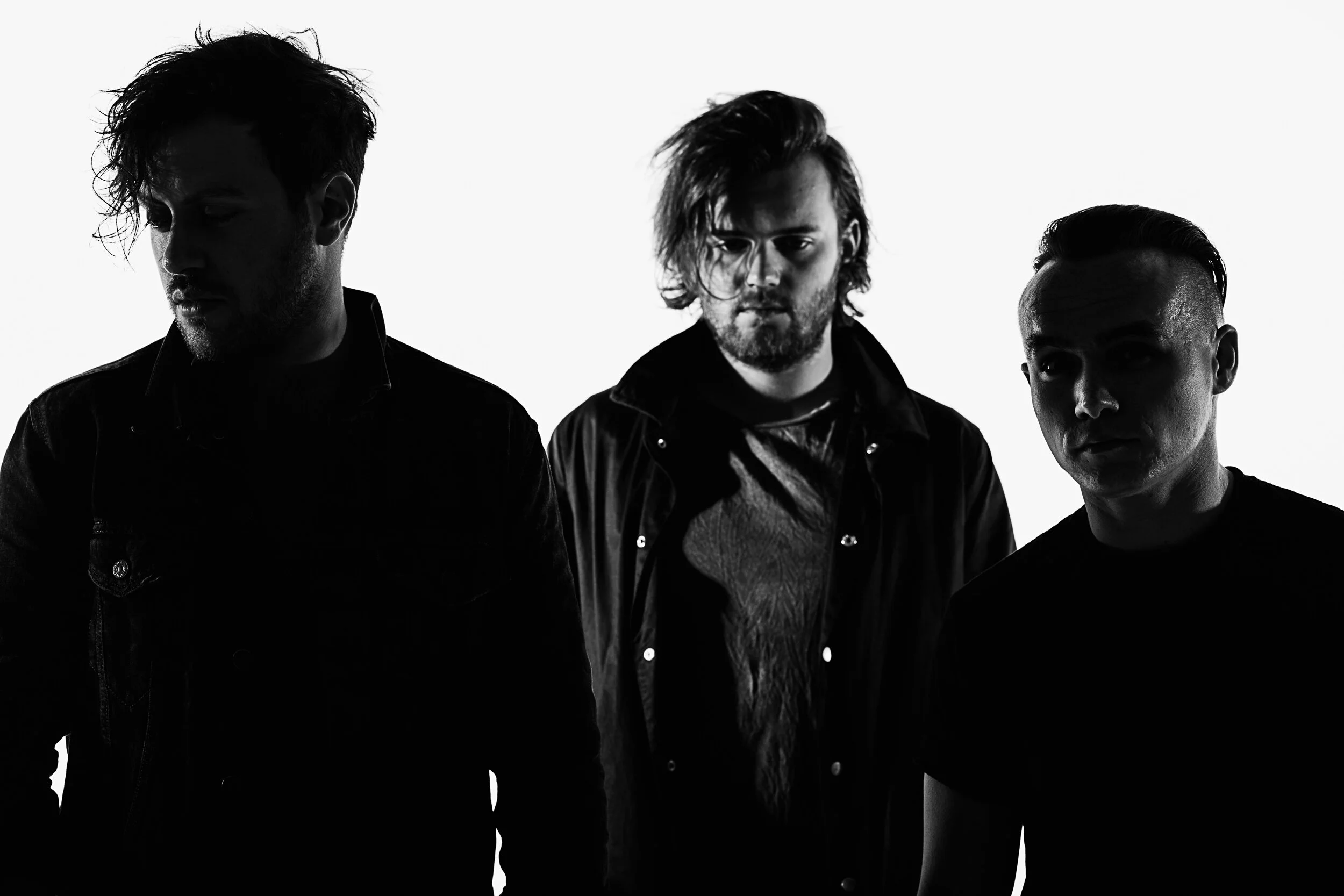 Black-and-white photo of three men with edgy hairstyles looking downward or to the side, dressed in dark clothing, against a bright background.