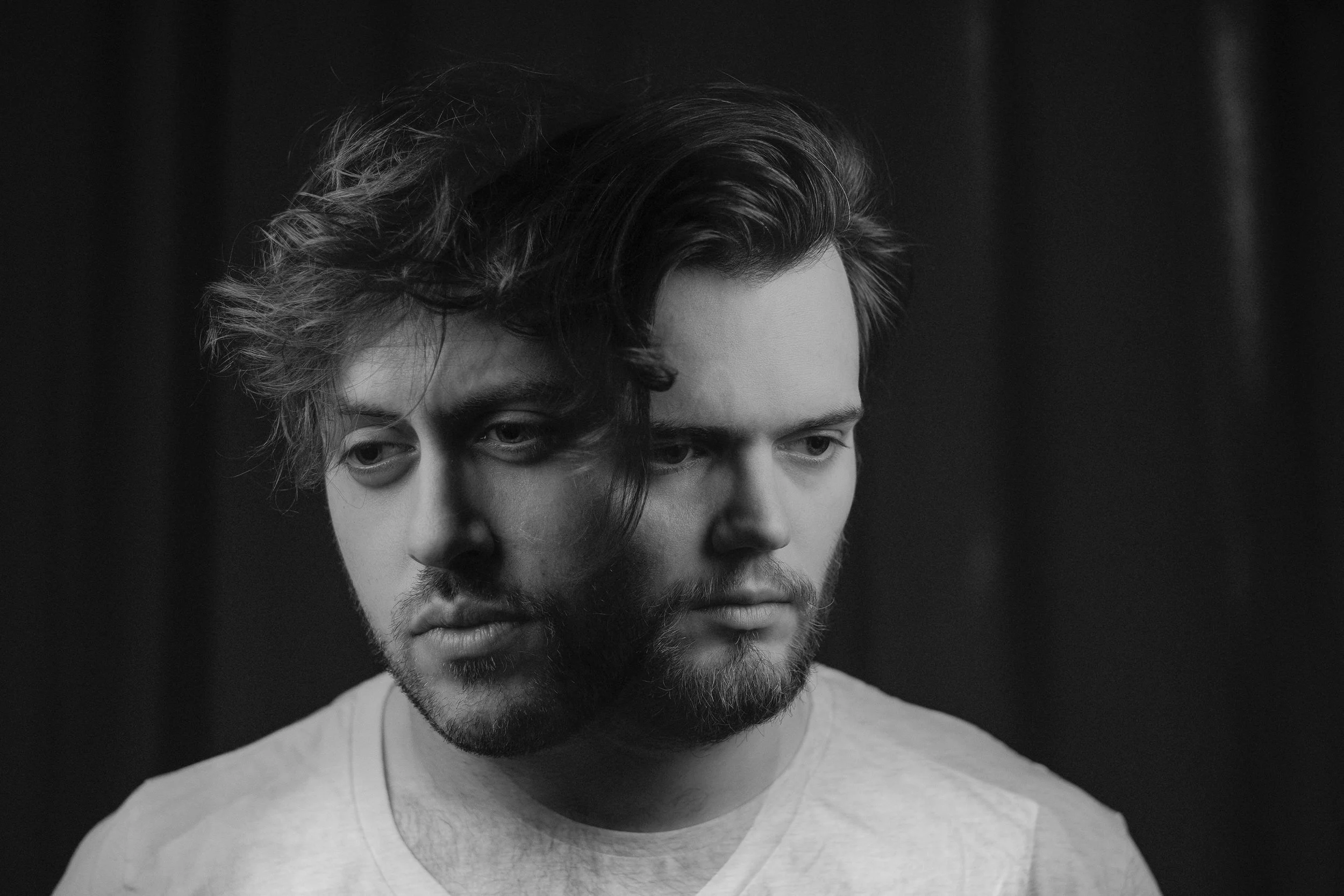 Black and white close-up photograph of a young man with wavy hair and a beard, looking down with a serious expression.