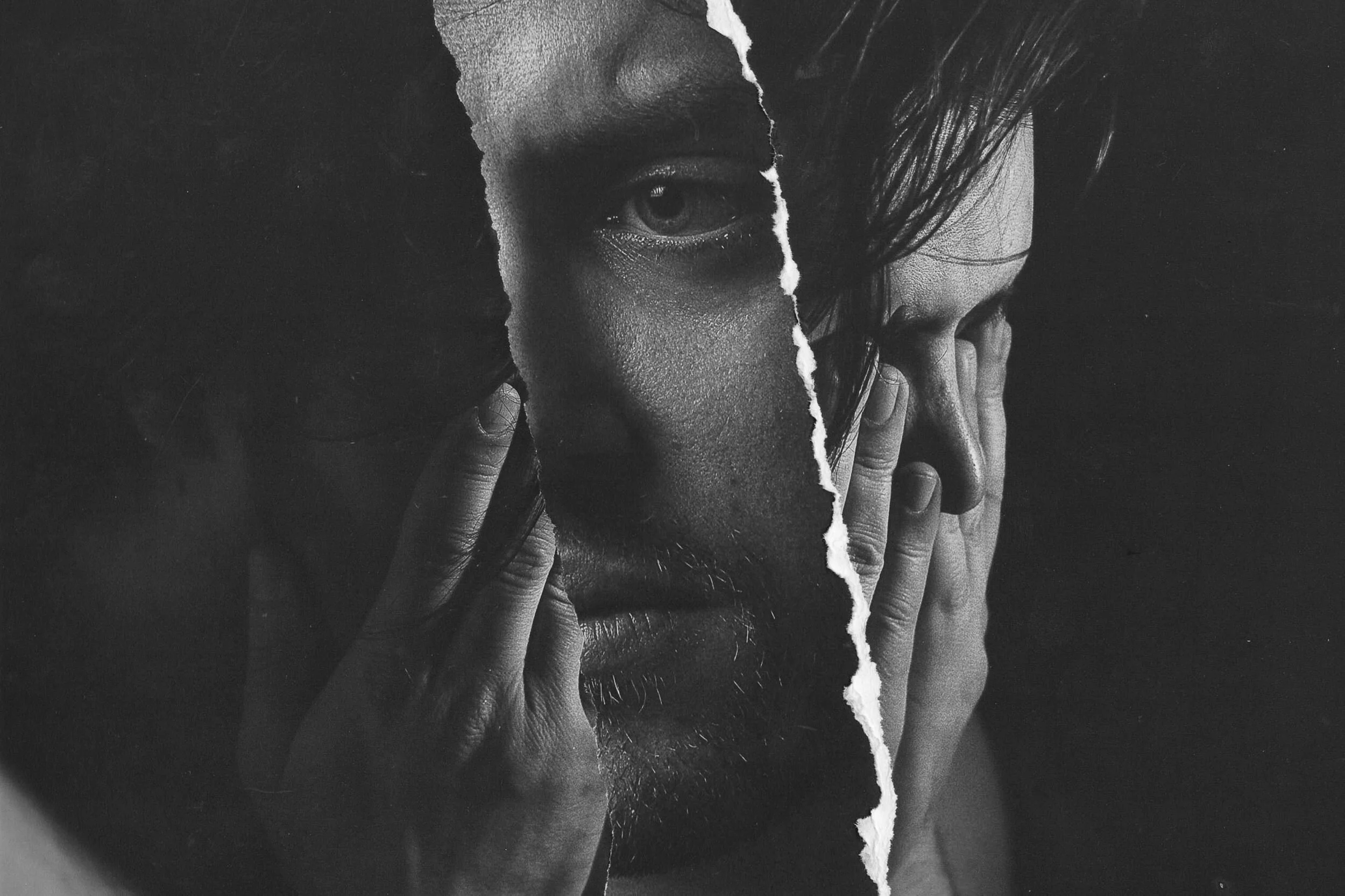 A black and white photograph of a person's face, partially covered by a torn paper with part of the face exposed, highlighting the eye, nose, and mouth, with hand gestures near the face.