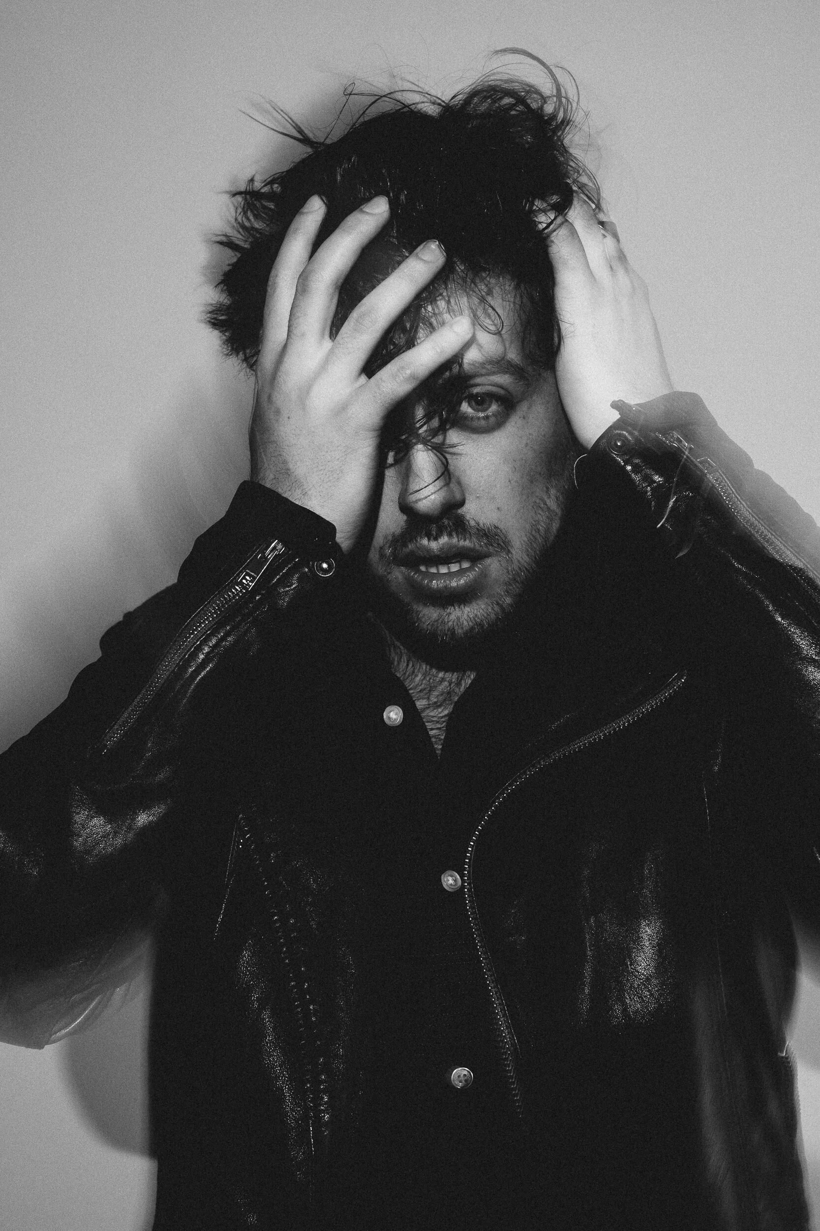 A person with dark hair and a beard wearing a black leather jacket, touching their head and face with both hands, looking into the camera with a serious expression.