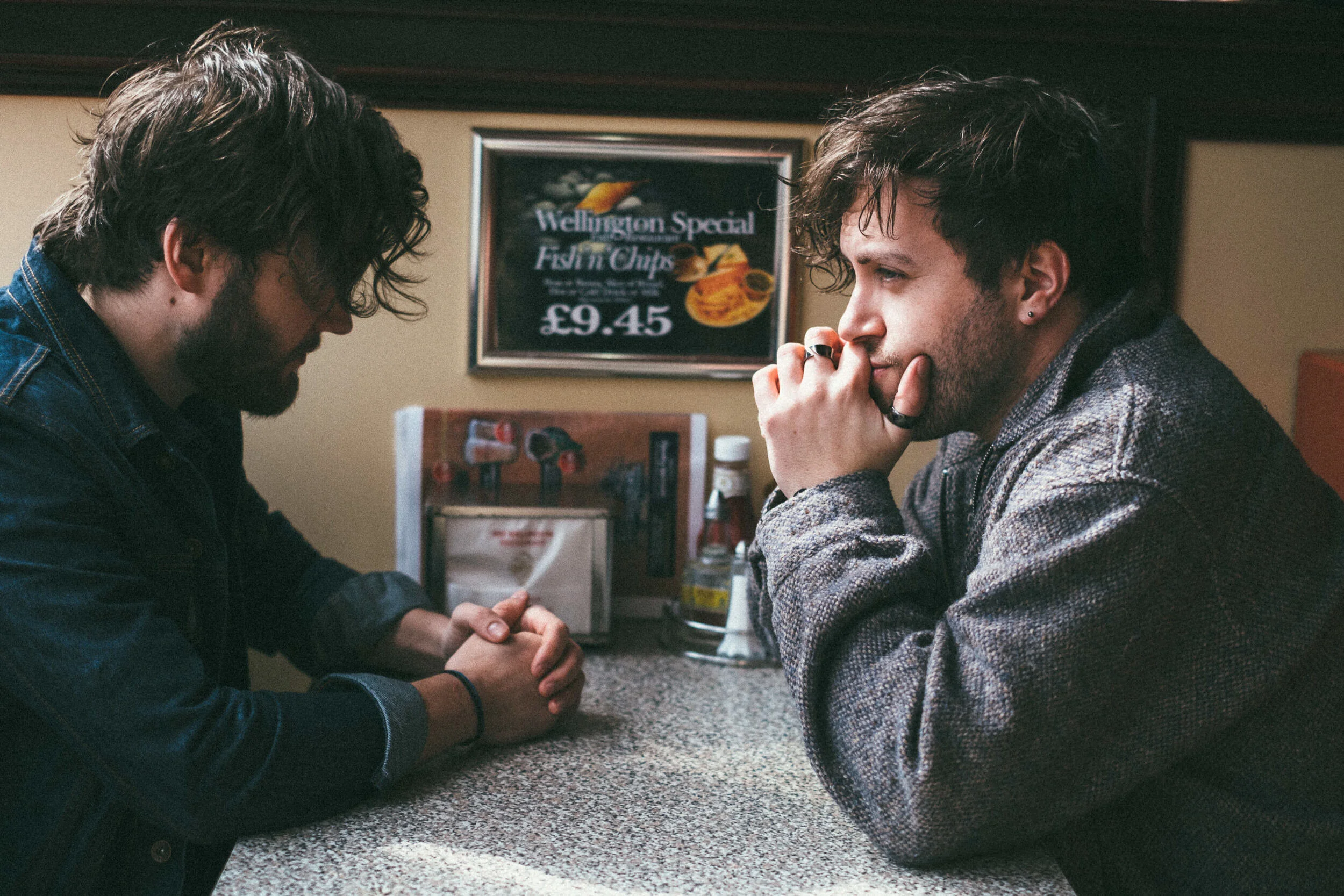 Two young men with messy hair and beards, sitting at a table in a casual setting, engaged in a serious conversation. They are holding hands and looking at each other, with one resting his chin on his hand. Behind them, a sign advertising fish and chi