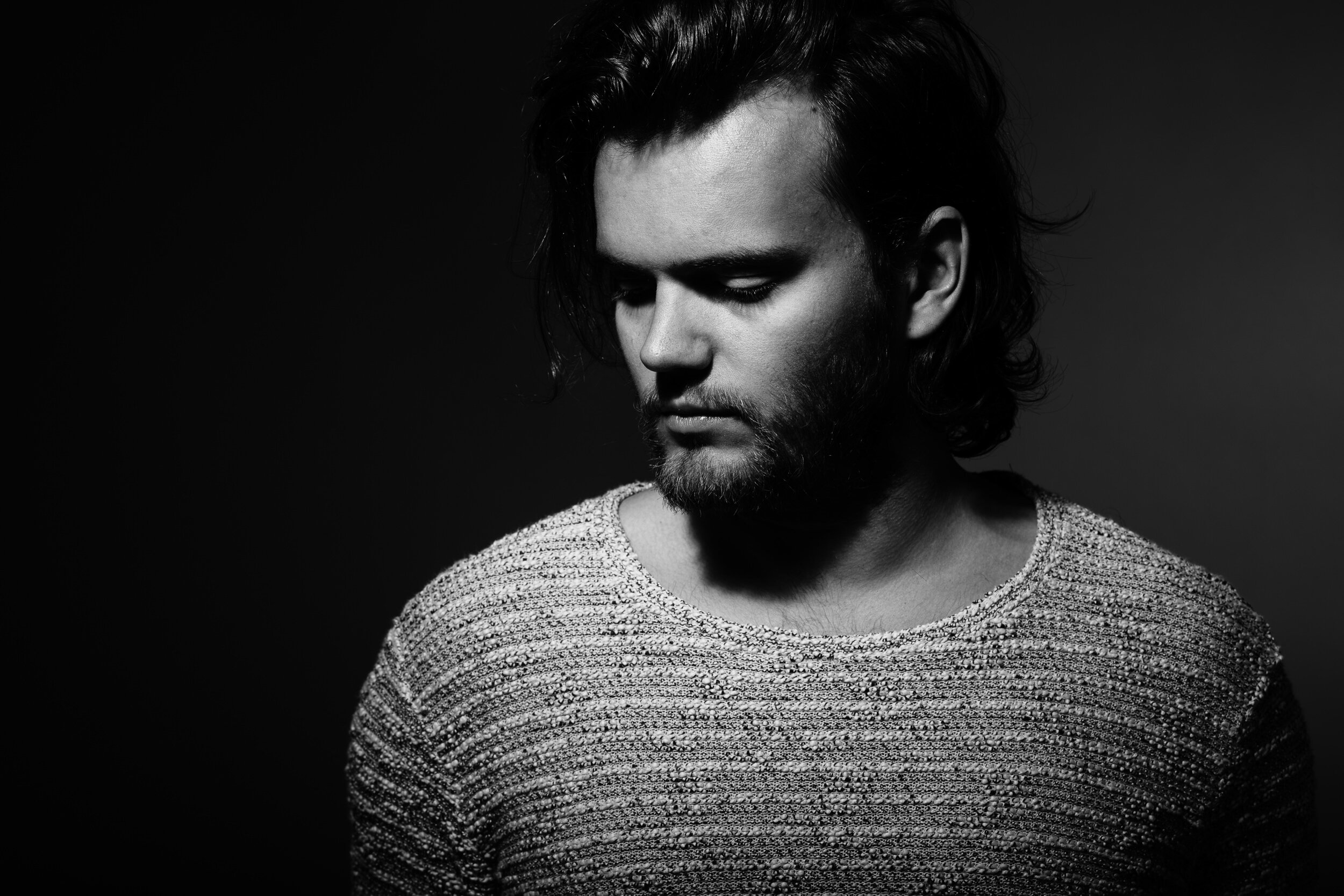 Black and white portrait of a young man with medium-length wavy hair, looking down, wearing a textured sweater.