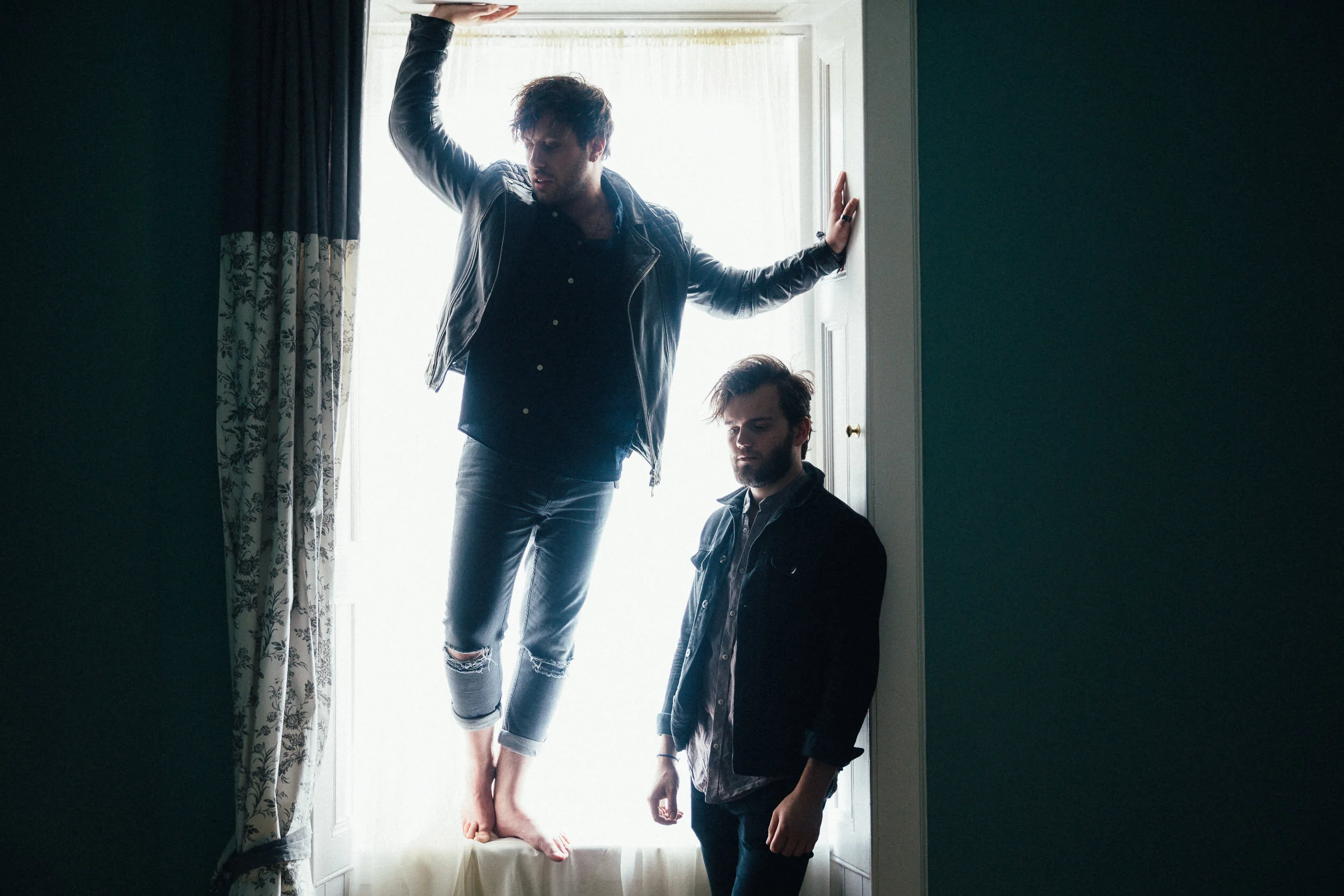 Two men in a room near a window with curtains. One man is standing with his arm extended, holding onto the wall, with his other arm raised and leaning against the wall. The other man is sitting or kneeling near the floor, with a serious expression.
