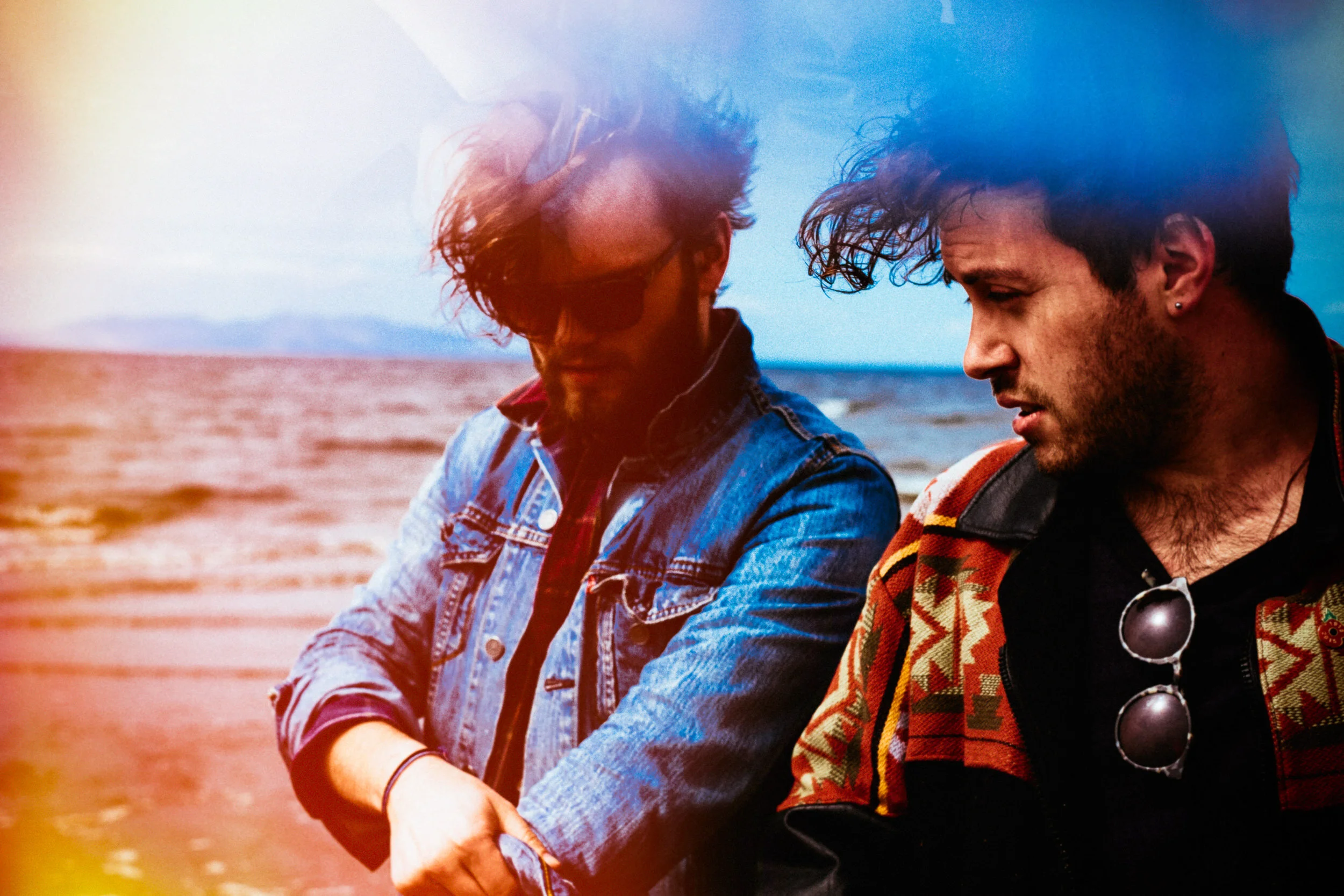 Two young men sitting outdoors near water, one wearing sunglasses and a denim jacket, the other with tousled hair and a patterned scarf, both looking down.