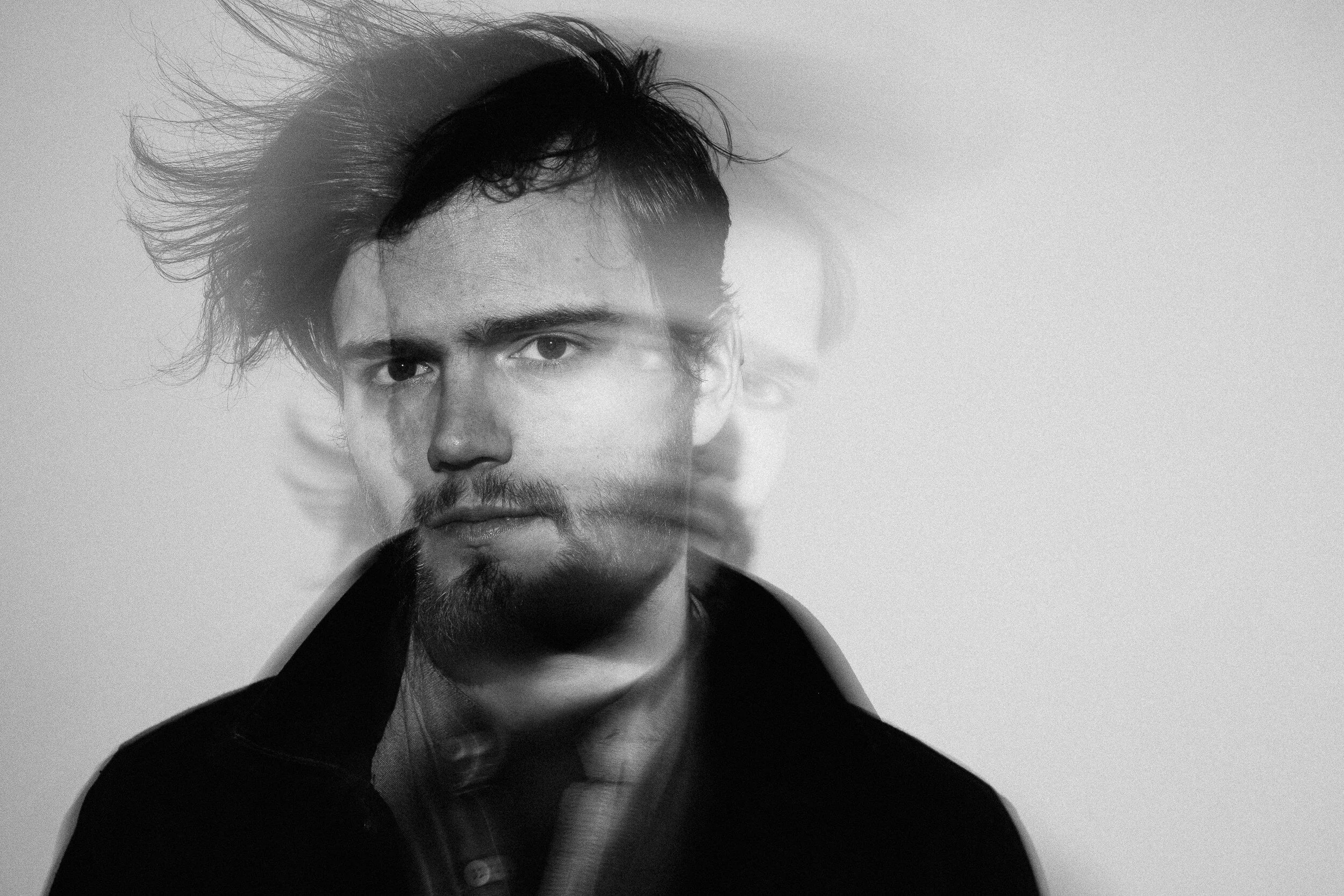 A black and white portrait of a man with dark messy hair, wearing a dark jacket over a shirt, against a plain light background.