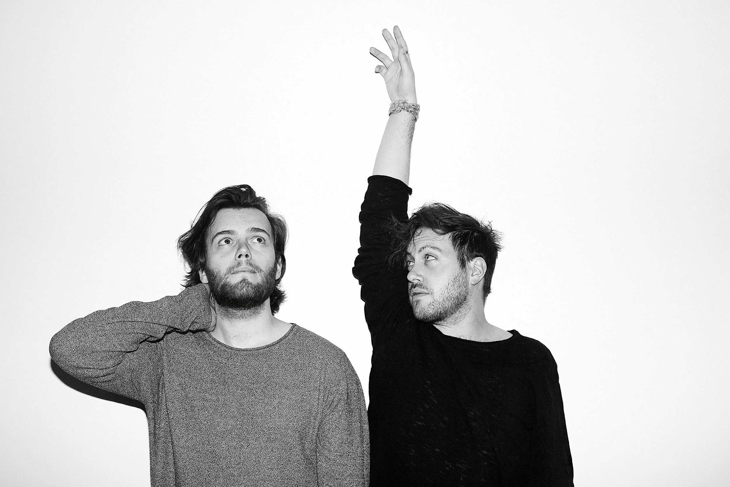 Two young men with beards and tousled hair, one with a sweater and the other with a dark shirt, standing close together against a plain background. The man on the right is raising his arm, with his hand positioned above the other man's head.