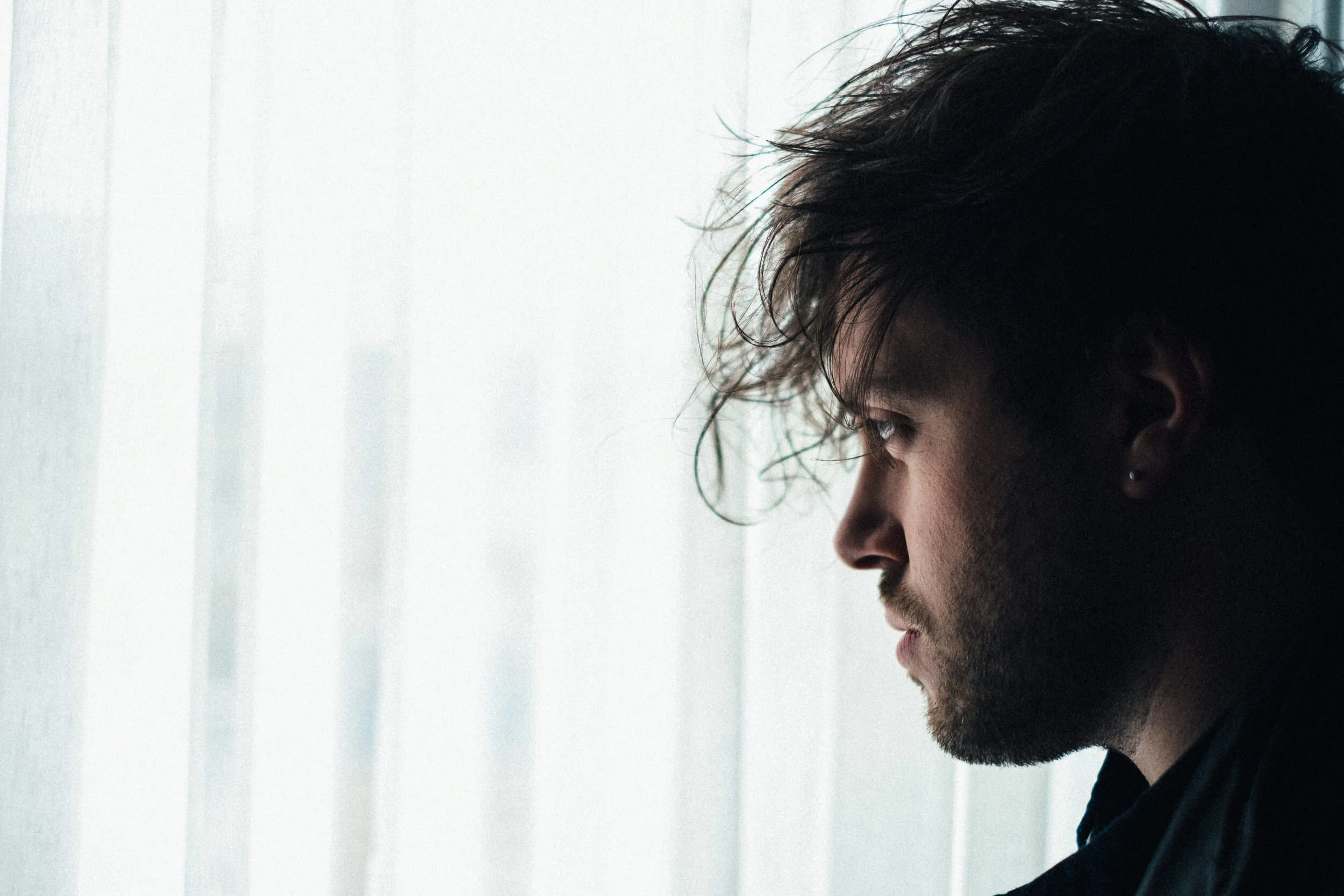 Profile of young man with dark hair and beard, looking downward, with light door or wall in background.
