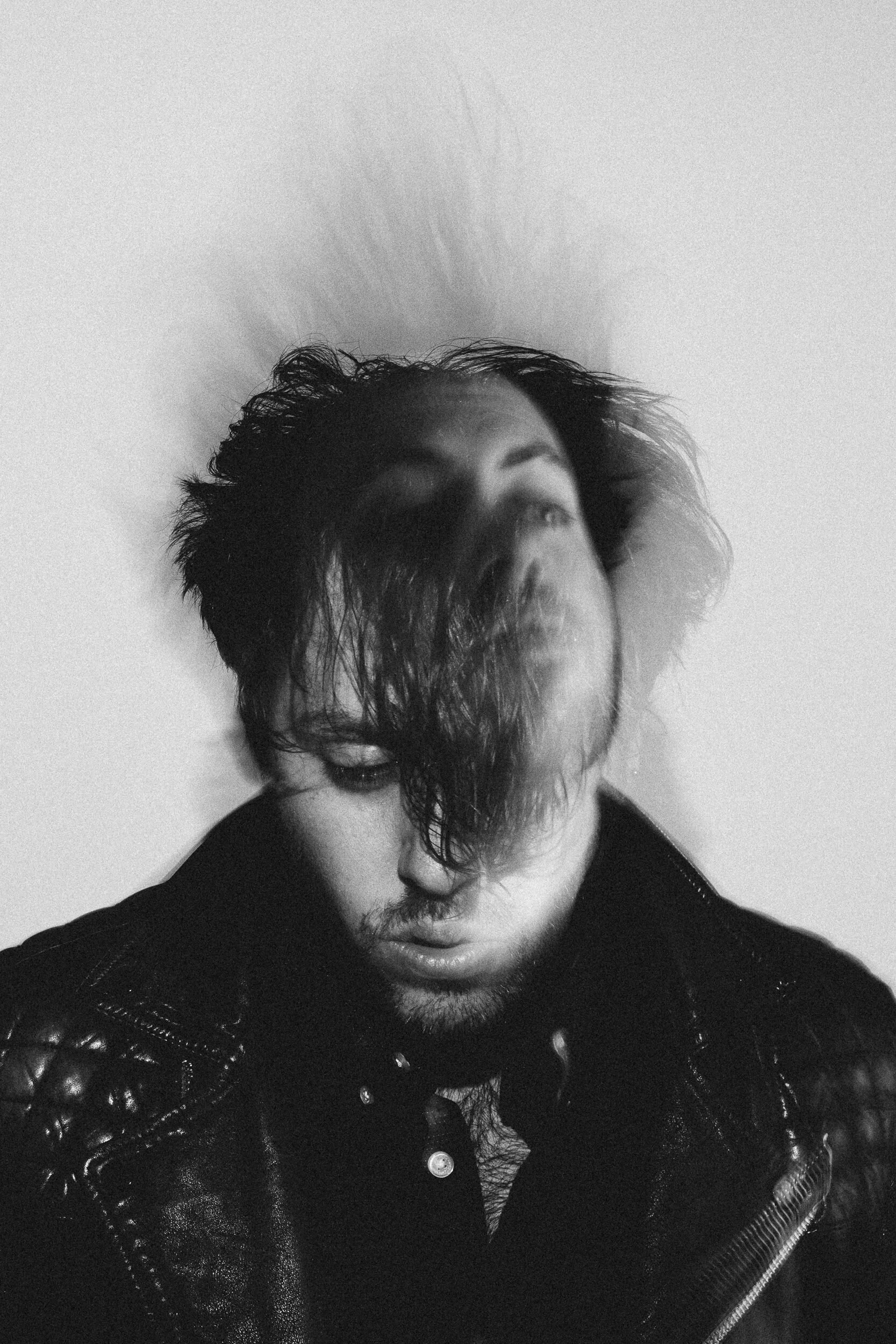Black-and-white photo of a man with curly hair and a beard, wearing a black leather jacket, looking downward.