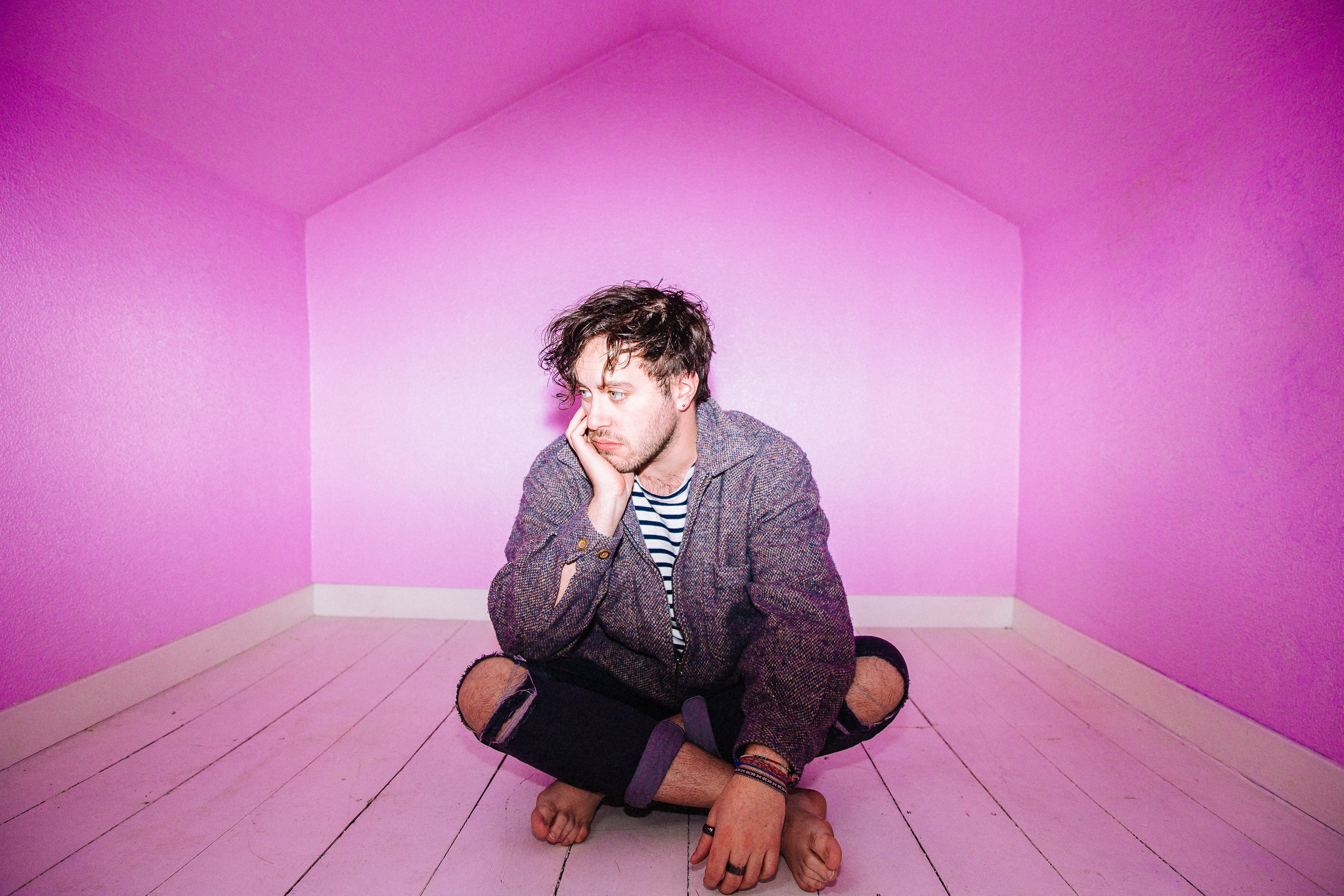 A young man with curly dark hair sitting on a pink wooden floor in a corner with pink walls, resting his chin on his hand, wearing a striped shirt and a textured blazer.