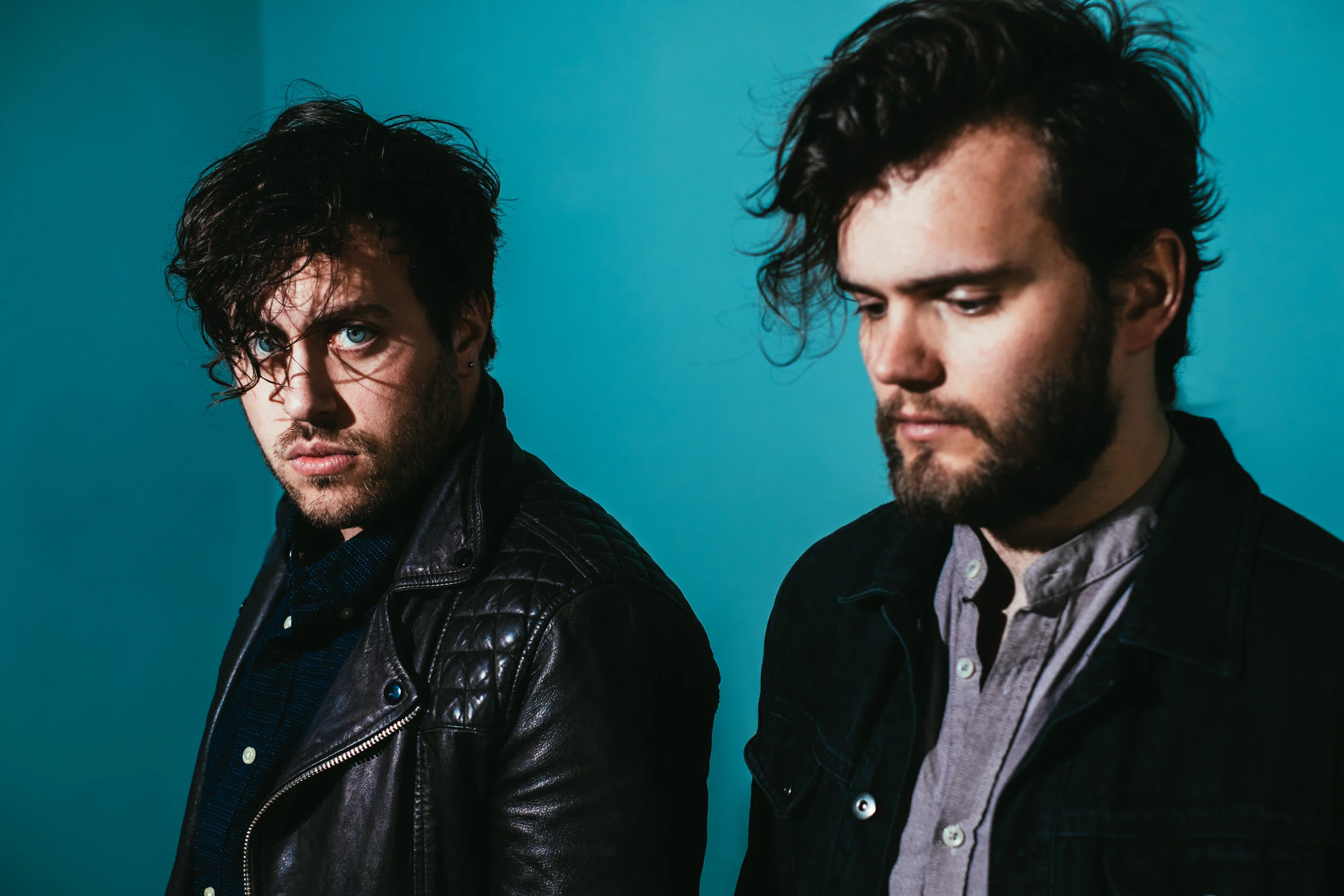 Two young men with dark, wavy hair and beards, dressed in dark jackets and shirts, standing against a teal background. The man on the left has bright blue eyes and is staring intensely at the camera, while the man on the right has his eyes cast downw