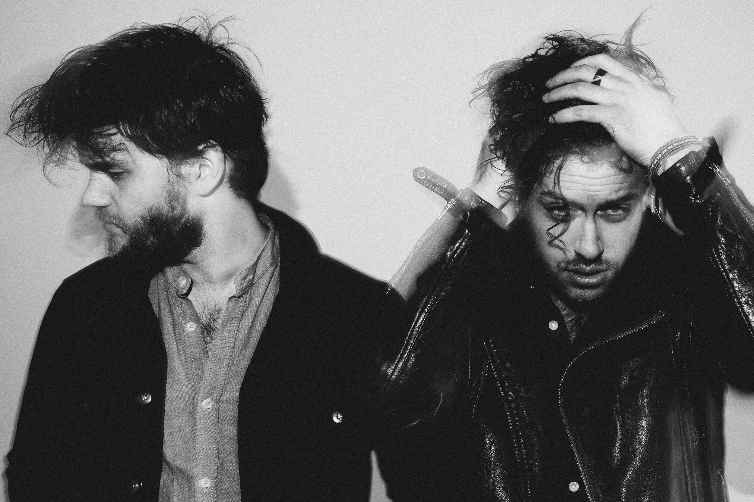 Black and white photo of two men with dark, messy hair and facial hair. The man on the left is looking down and to the left, wearing a collared shirt and a dark jacket. The man on the right is touching his head with both hands, wearing multiple brace
