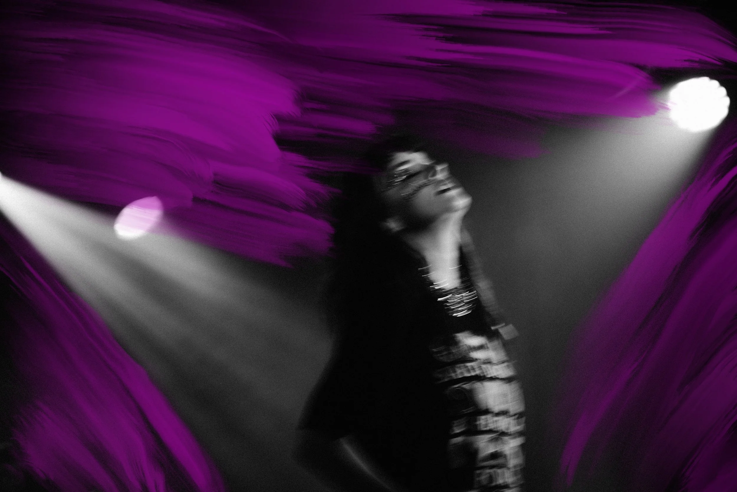 A motion blur, black and white image of Lauren Mayberry performing a concert. She is alone on stage under a spotlight. Purple oil paint has been added to the image.