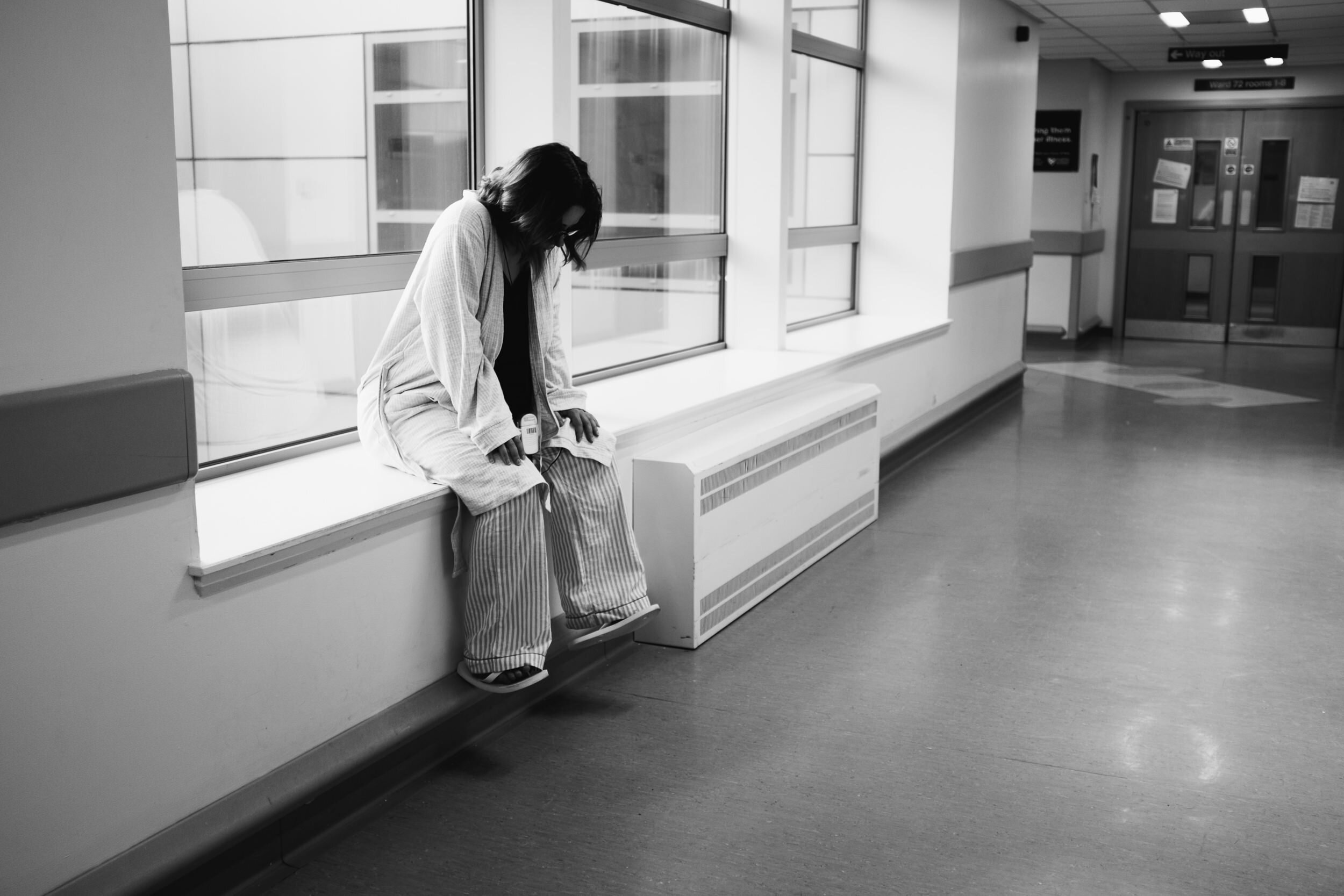 A woman sitting alone on a hospital waiting room bench, looking down with her head resting on her hand, in a black and white photo.