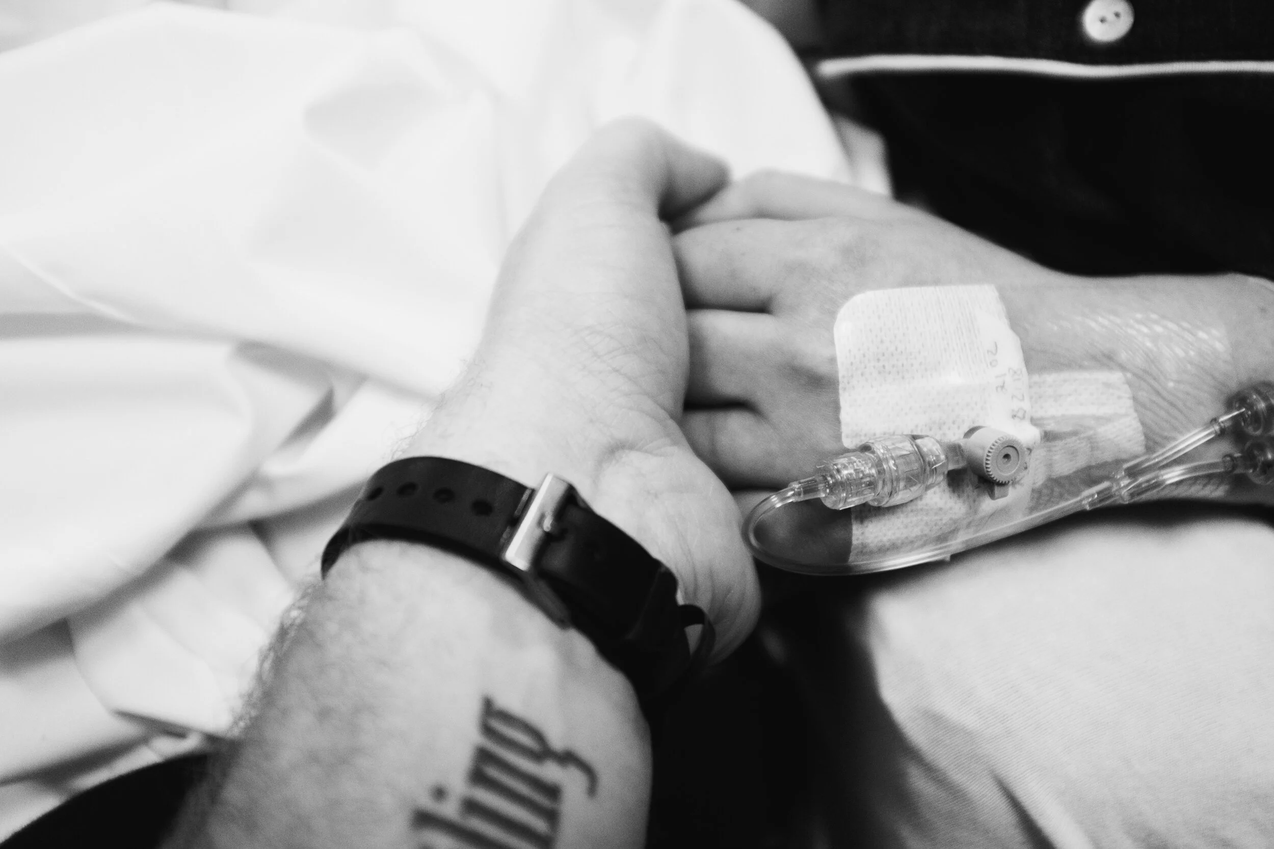 Close-up of a person's arm with a hospital bracelet and intravenous (IV) line, holding another person's hand with medical tape on their wrist, in a hospital setting.