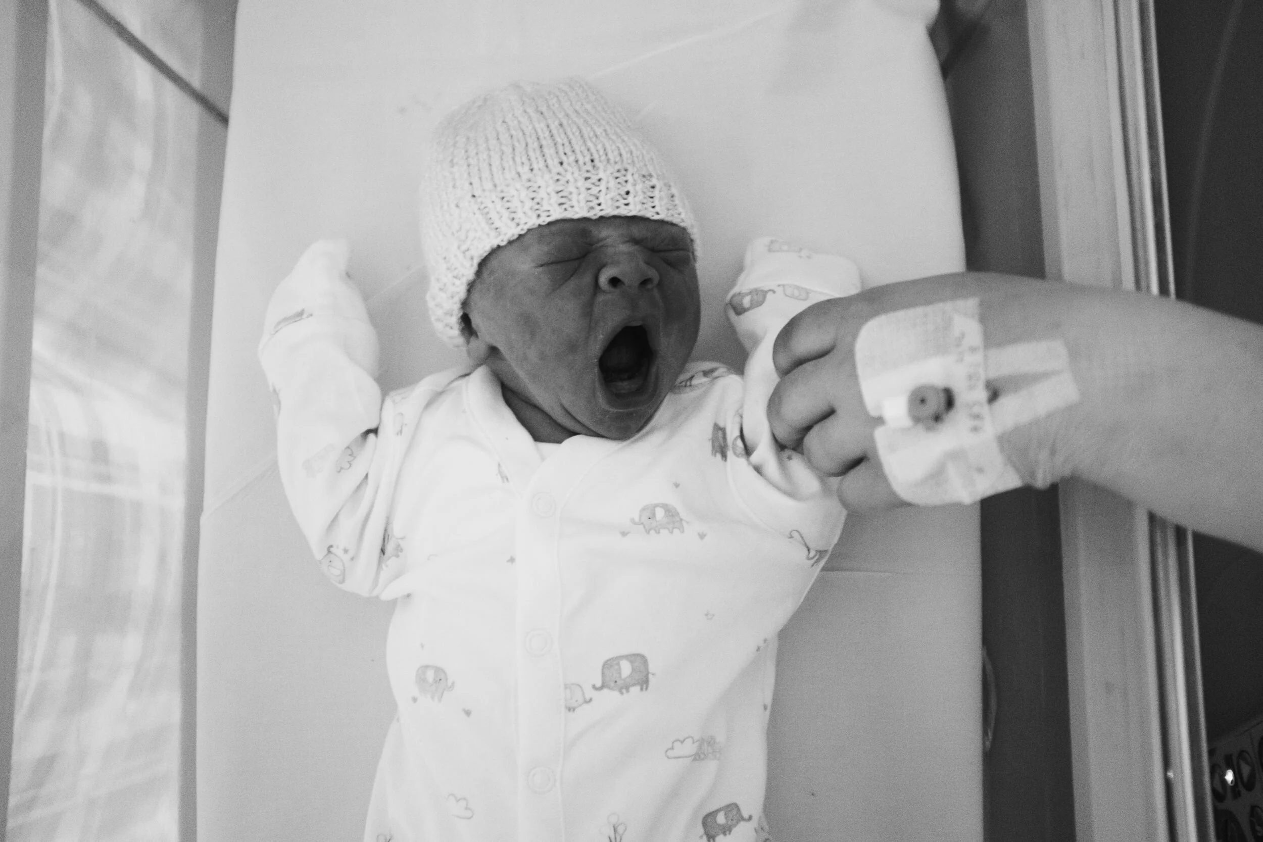 A newborn baby wearing a knitted hat and patterned hospital gown, yawning with its eyes closed, in a hospital bed.